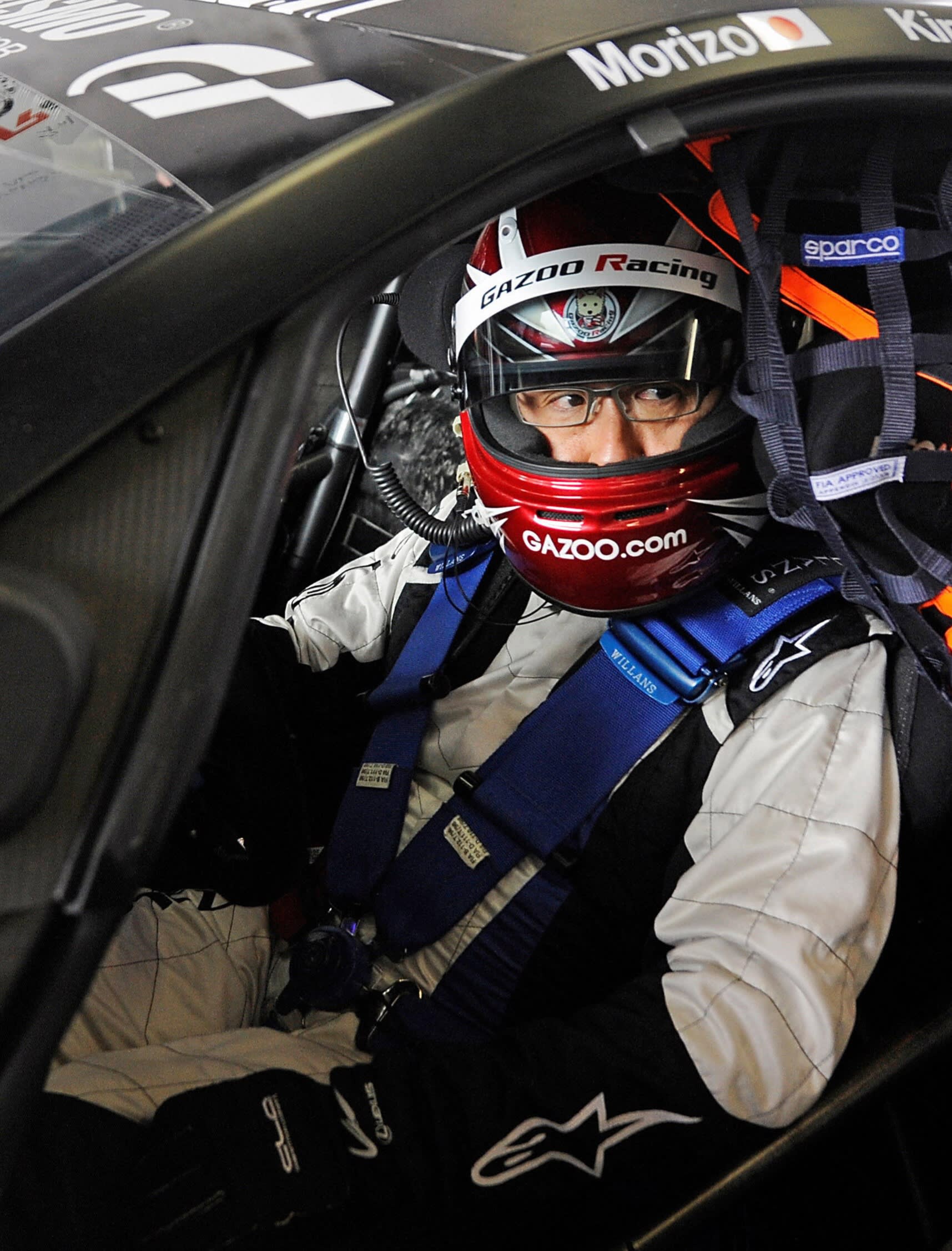A race car driver wearing a red Gazoo Racing helmet and white racing suit sits inside a car, strapped in with blue safety harnesses