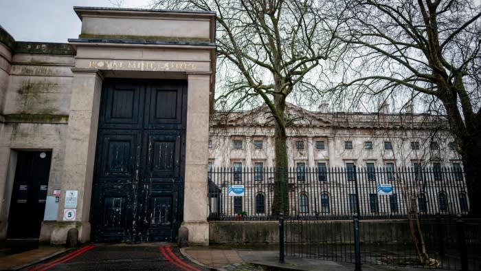 Entrance gate of Royal Mint Court with large black doors, weathered stone walls, and a historic building in the background