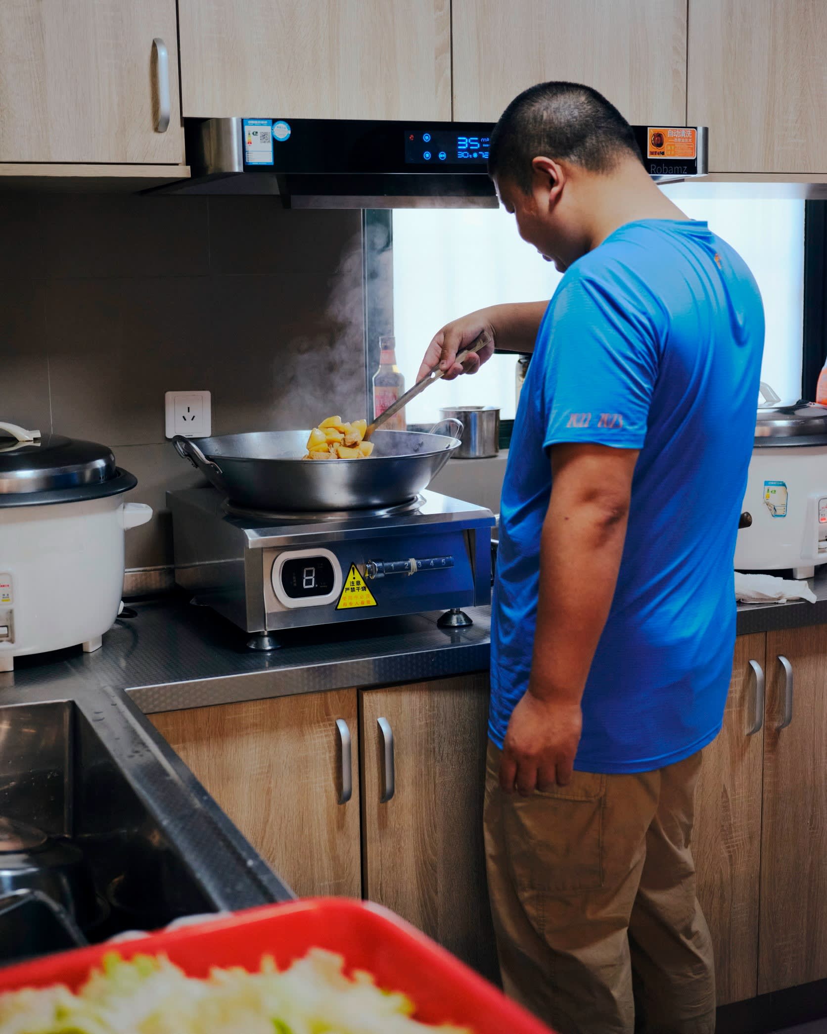 A man in a blue T-shirt stands at a counter, stirring food in a pan