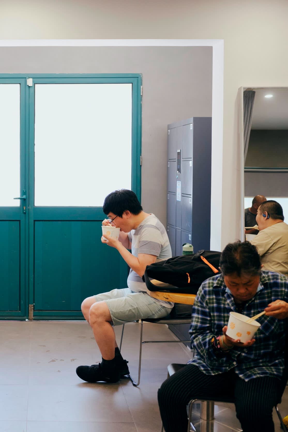 Men sit on chairs in the lobby of an office like space, holding up bowls of food which they are eating with chopsticks