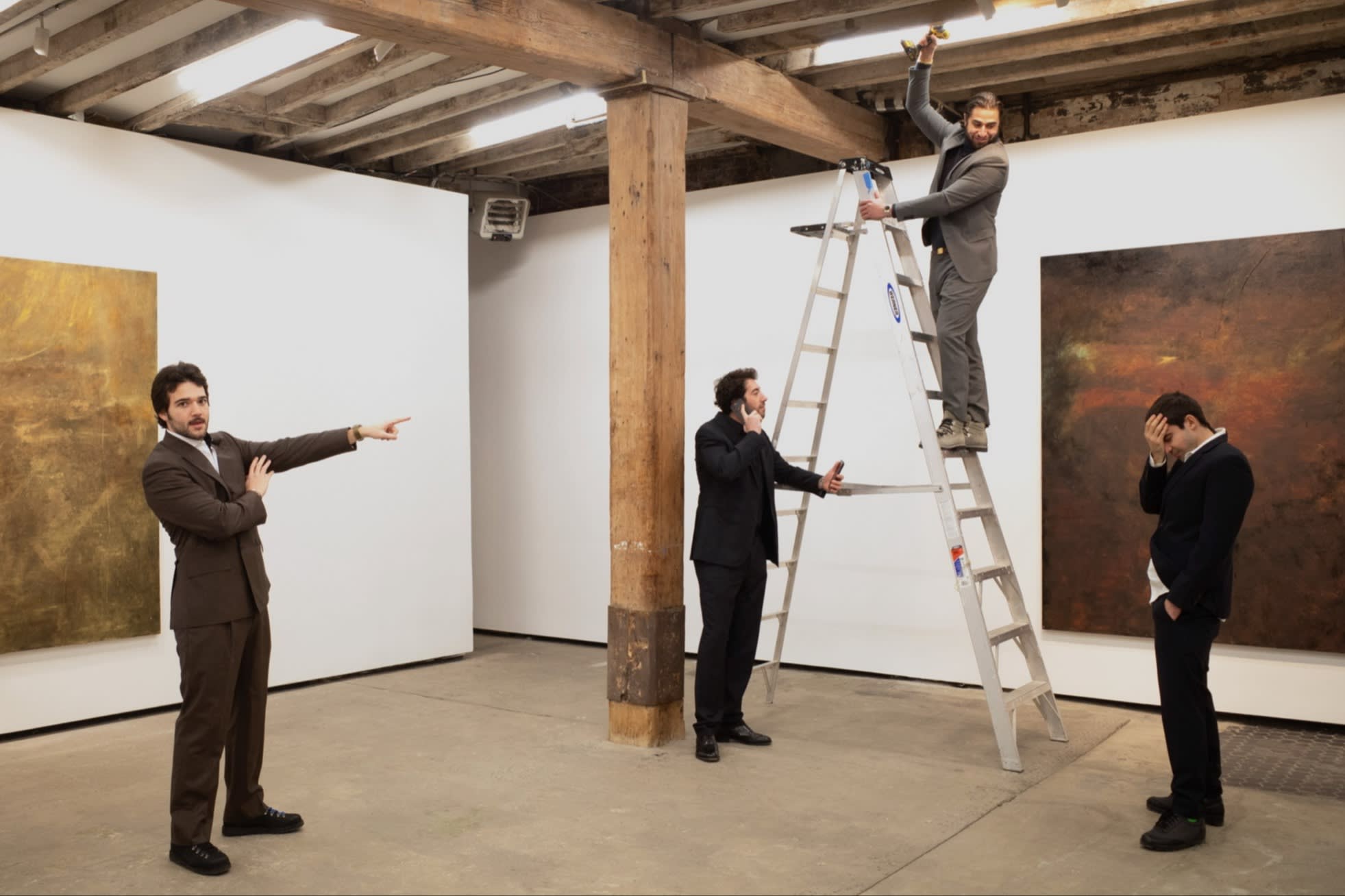 Four men wearing suits stand in an art gallery, adopting different poses; one is up a step ladder, reaching between beams in the ceiling