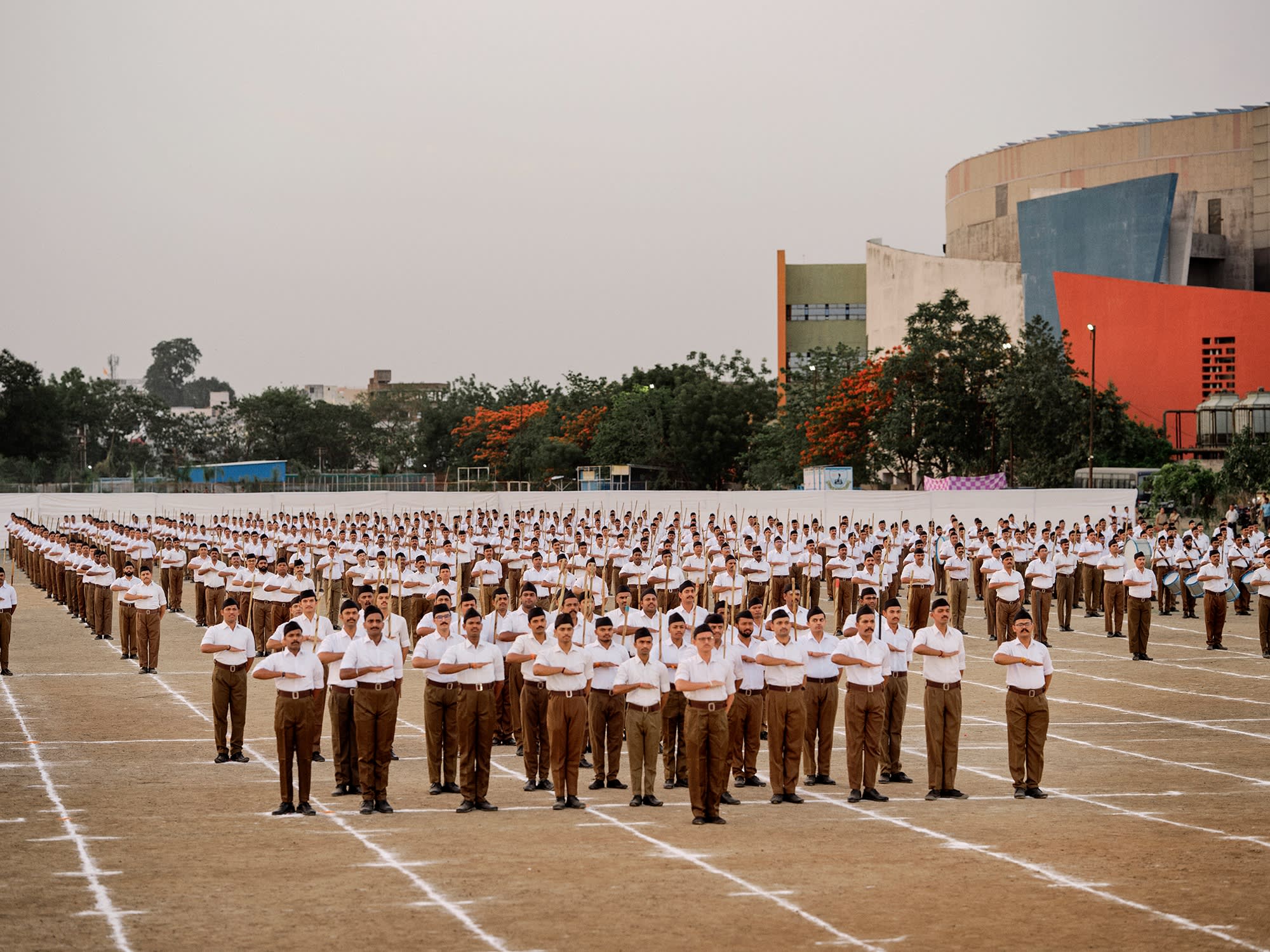 Rows of men in white shirts and brown trousers stand at attention in a large open field