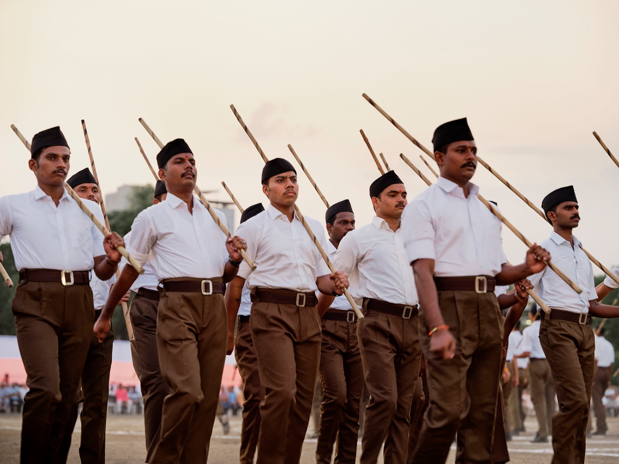 Uniformed men march shoulder to shoulder carrying sticks at a public gathering