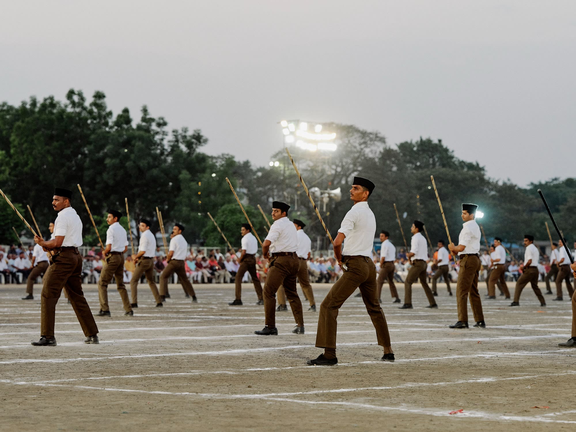 Group of men stands in formation holding sticks, with spectators in the background