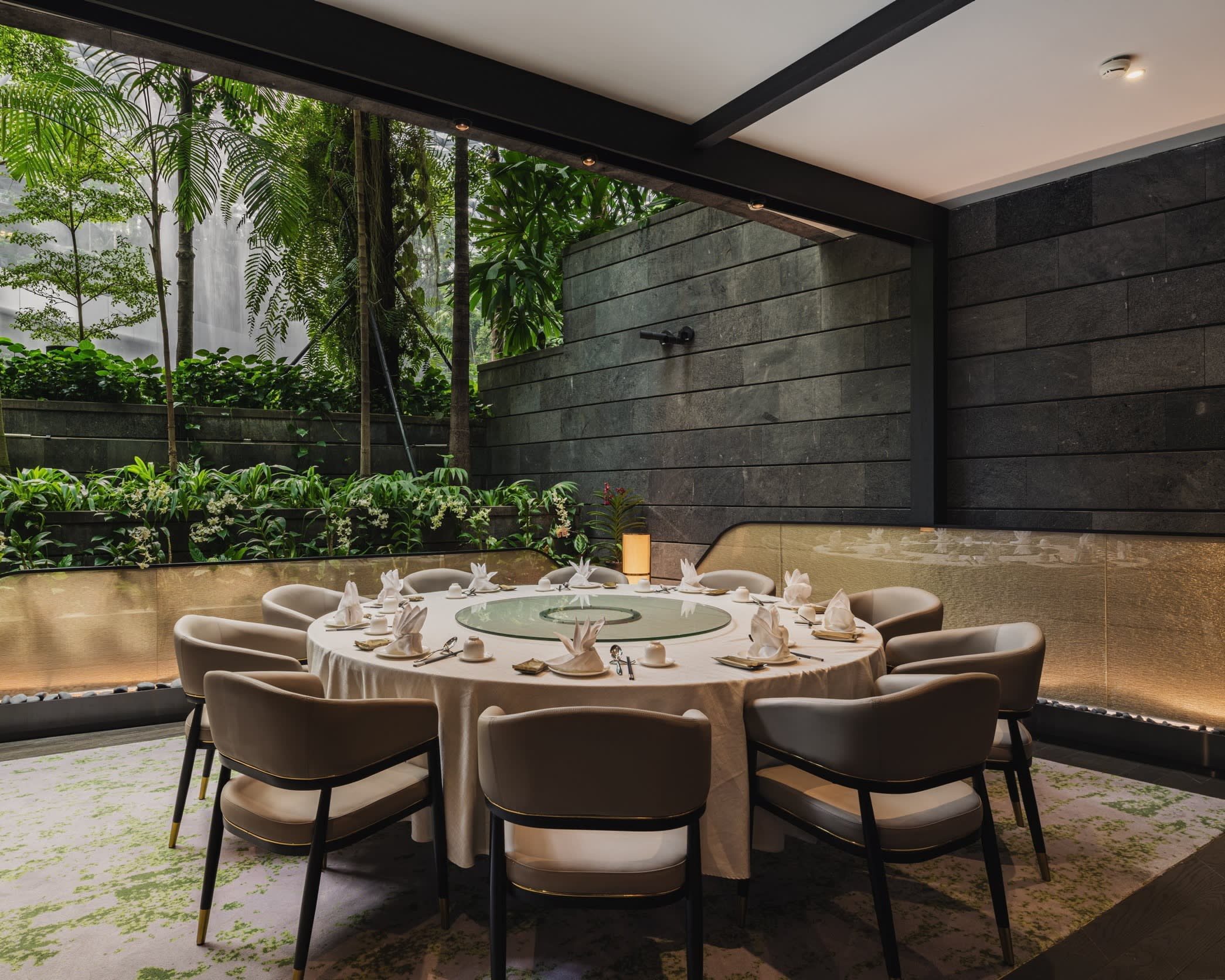 A circular dining table surrounded by ten chairs in the modernist dining room of a restaurant, bordering on to what looks like an outdoor area with plants and trees