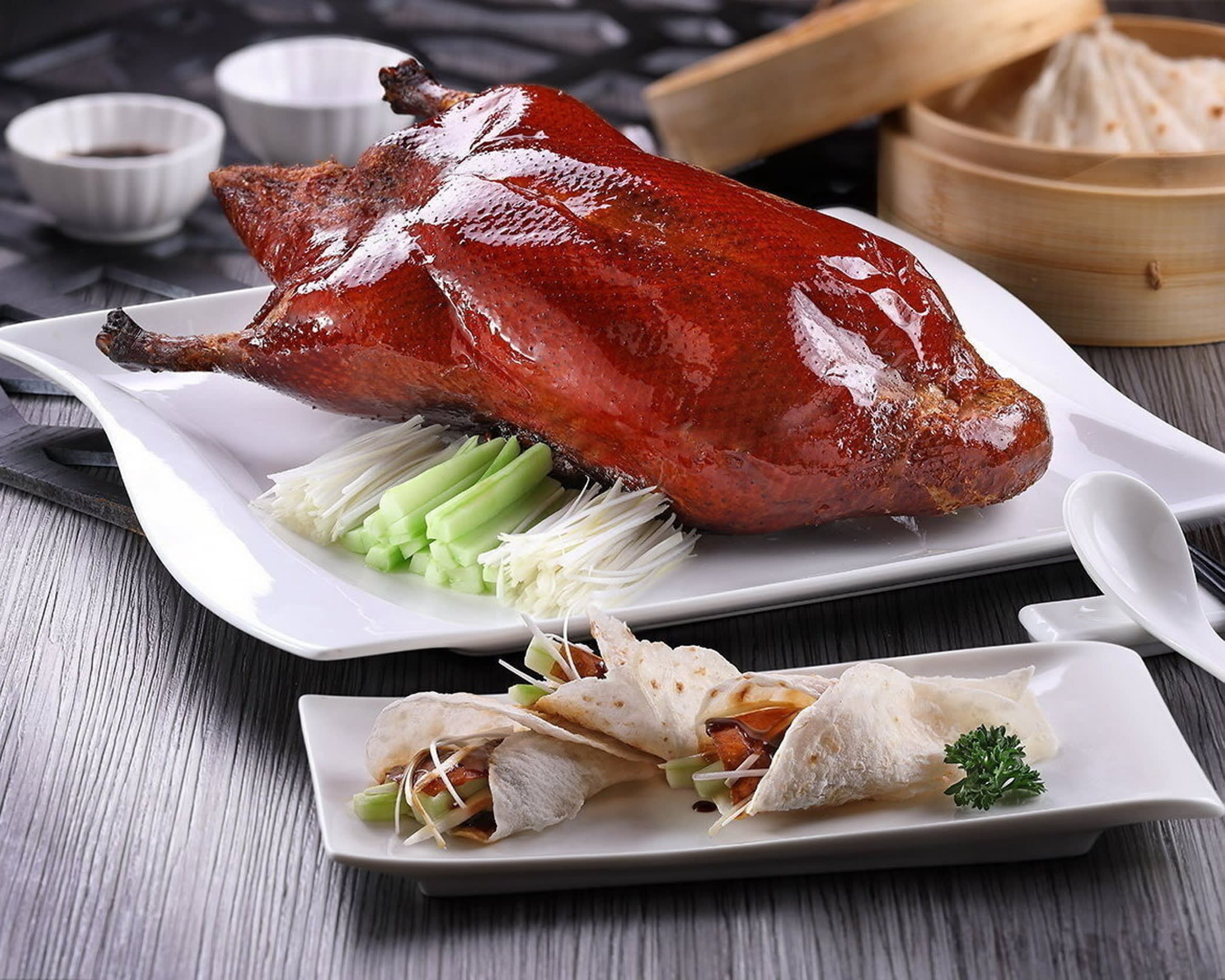 A whole duck, cooked Peking duck style, with glazed red coat, sits on a bed of chopped spring onions and chopped celery on a white plate, with spring rolls on another plate 