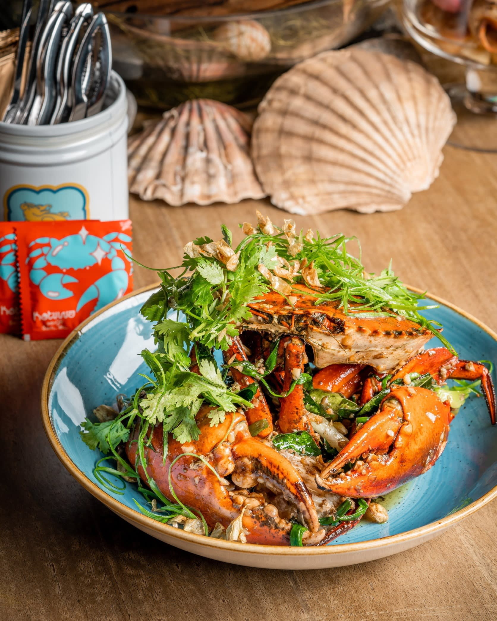 A spiced cooked crab dish on a plate, amidst a green salad. Around it on the table is a jar containing cutlery and several ornamental shells