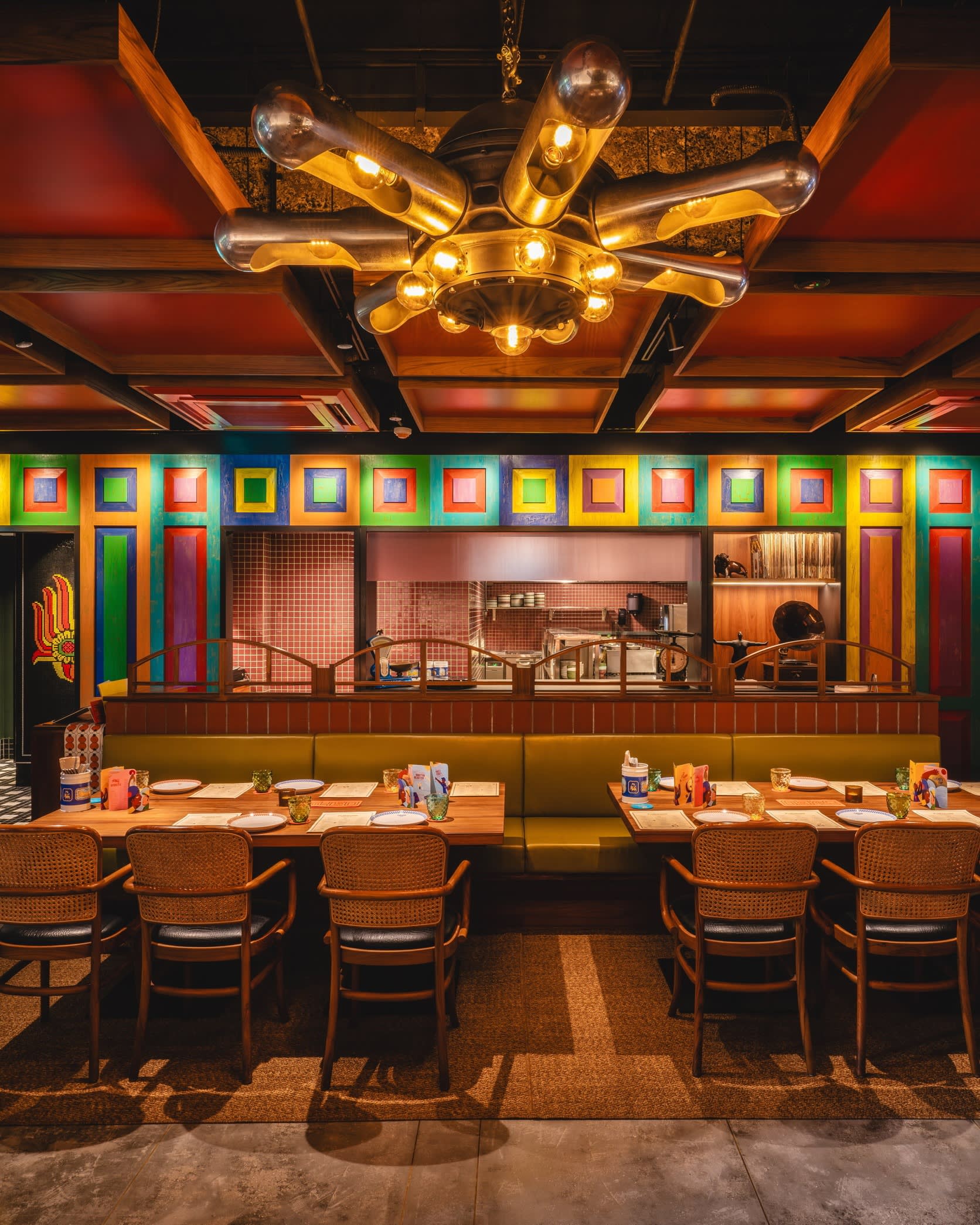 Colourful restaurant: in the background is the counter, with part of the kitchen visible behind it; in the foreground is a row of dining tables and chairs 