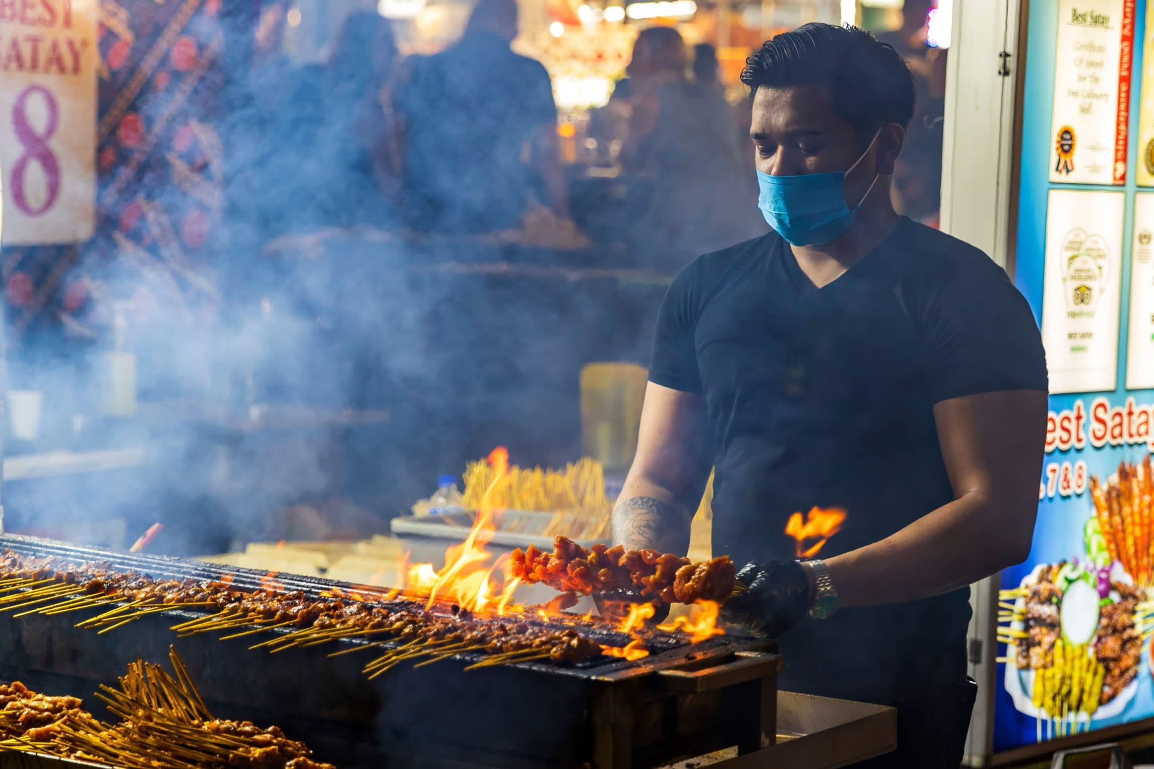 A man wearing a blue mask and a black T-shirt grills meat on skewers on a flaming outdoor grill at Lau Pa Sat hawker market in Singapore