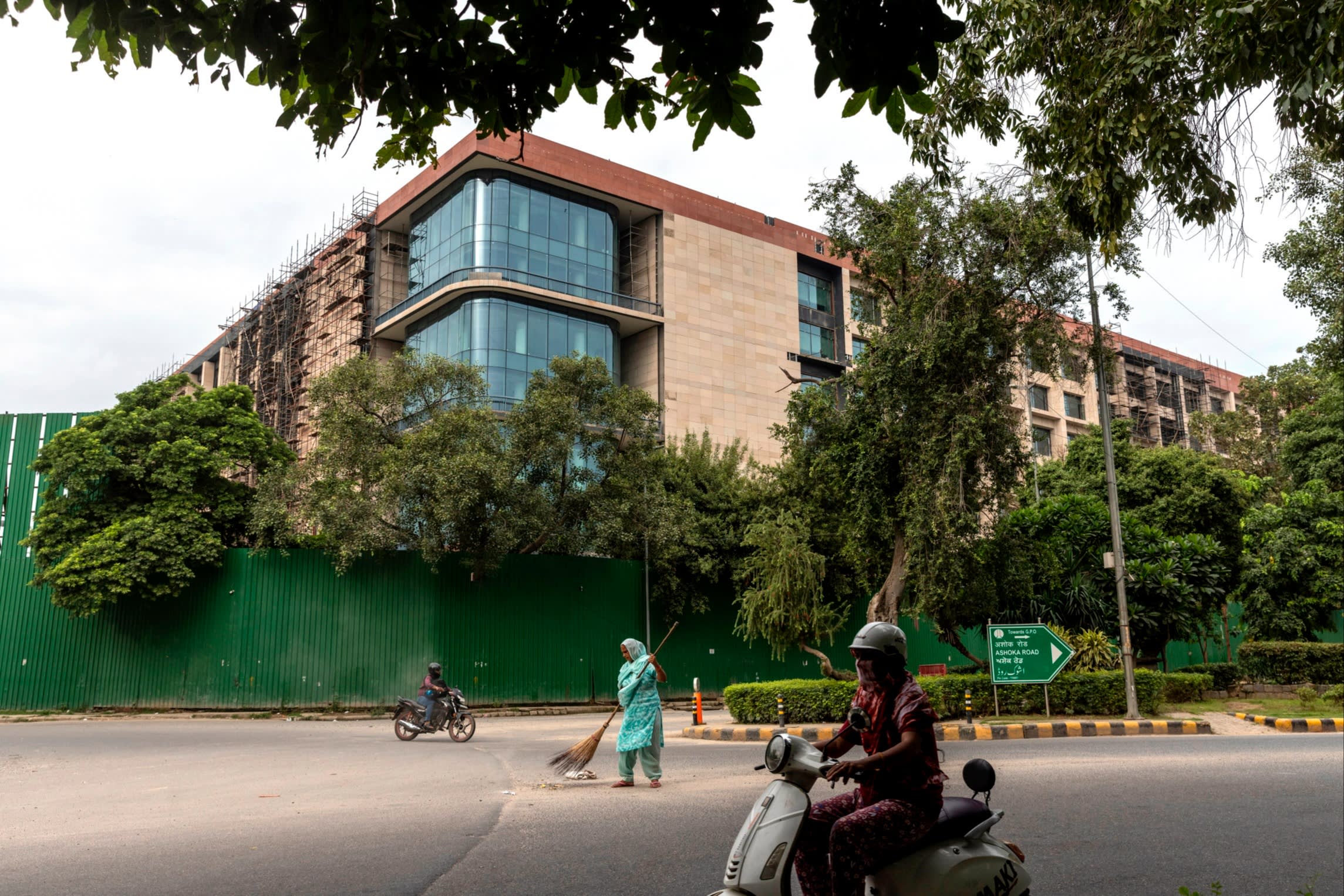 A person a moped drives past a large office building. Behind them, a woman is sweeping the road