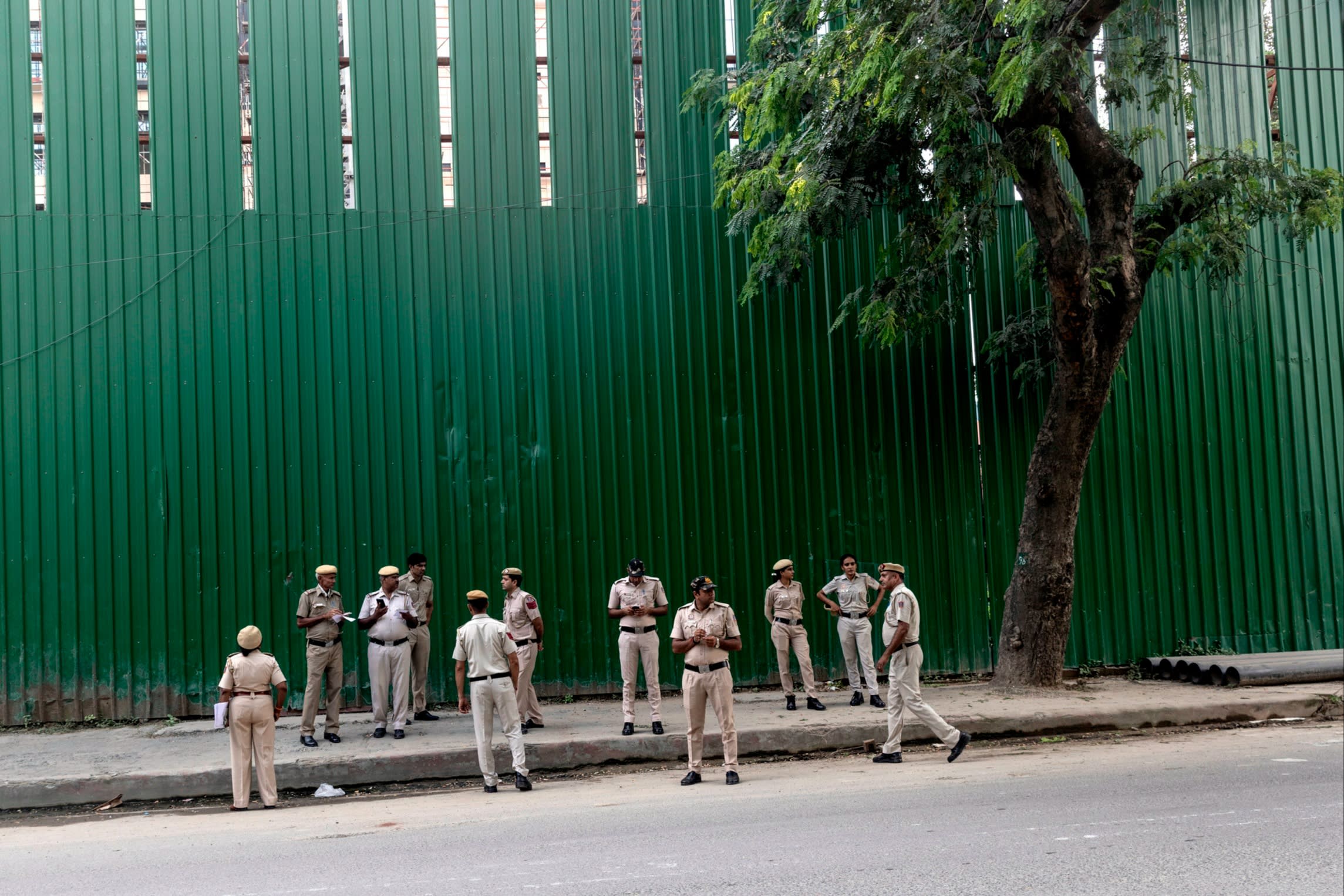 A group of uniformed officers stand in a street, in front of a tall green corrugated iron hoarding