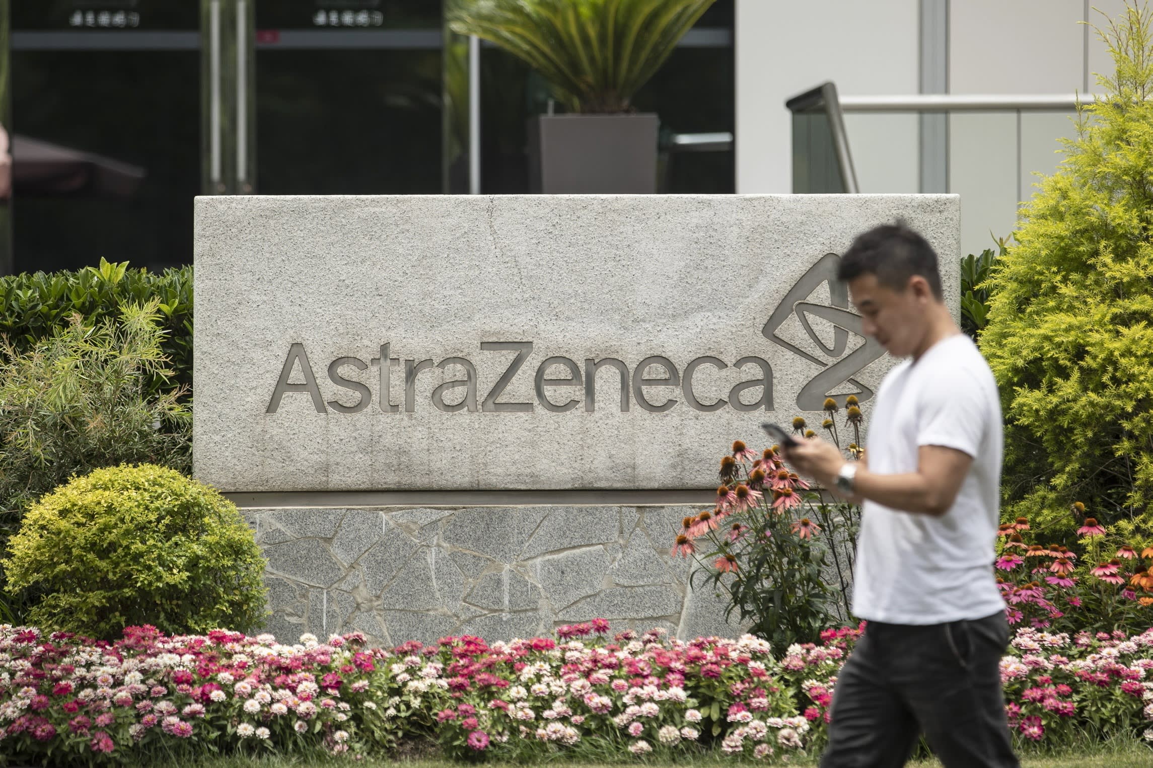A person walks past a large AstraZeneca sign surrounded by flowers outside a research facility.
