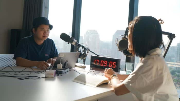Cheng Yanliang sits at a table with podcast equipment and a laptop, speaking into a microphone across from another person.