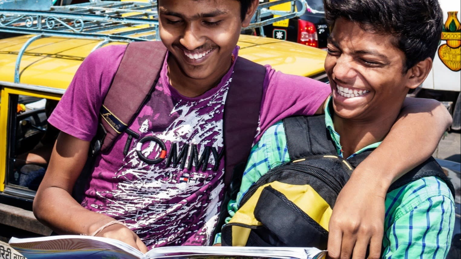 Two Indian teenagers embrace and laugh as they read a magazine together