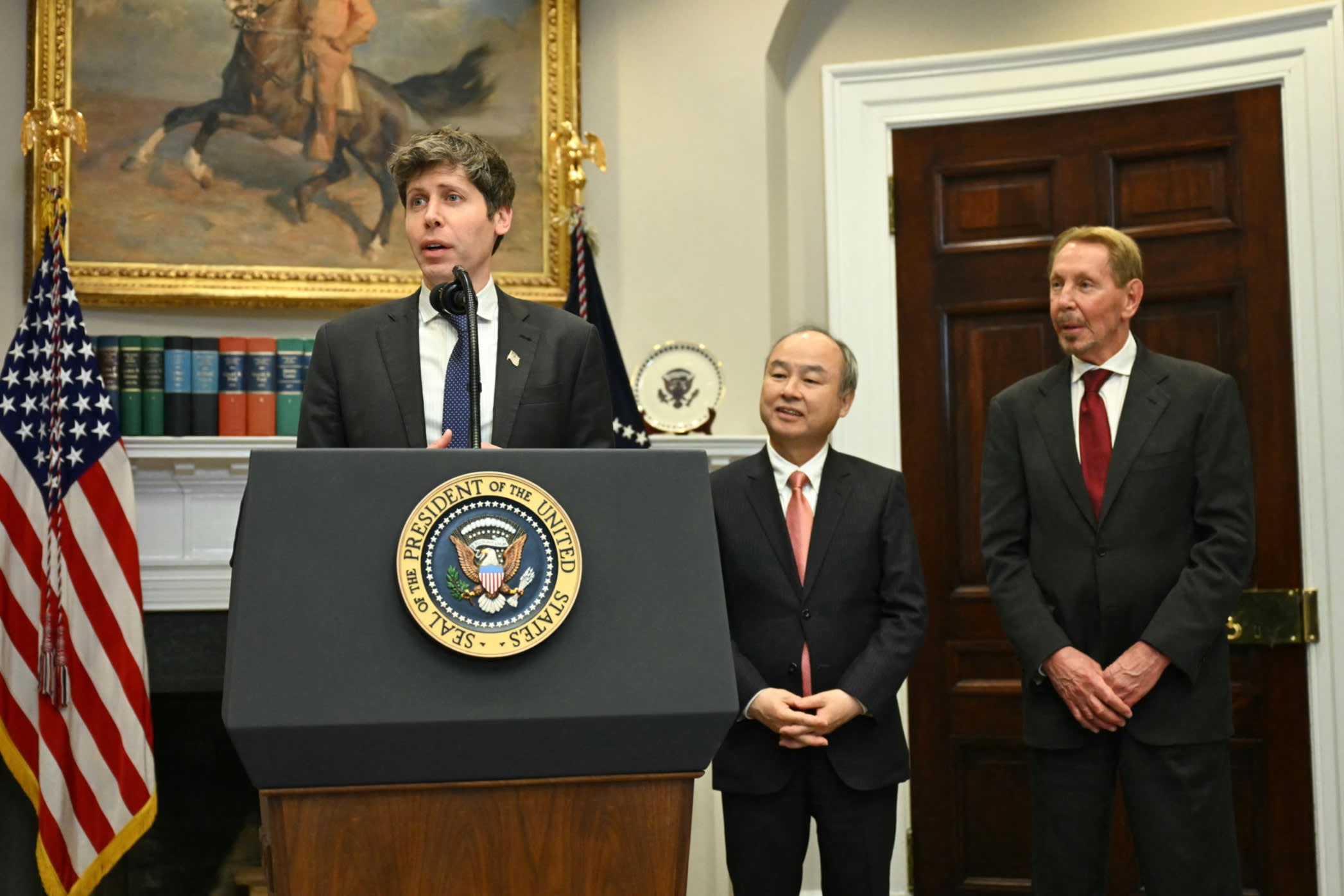 Sam Altman speaks at a podium while Masayoshi Son and Larry Ellison stand and listen beside him in the Roosevelt Room.
