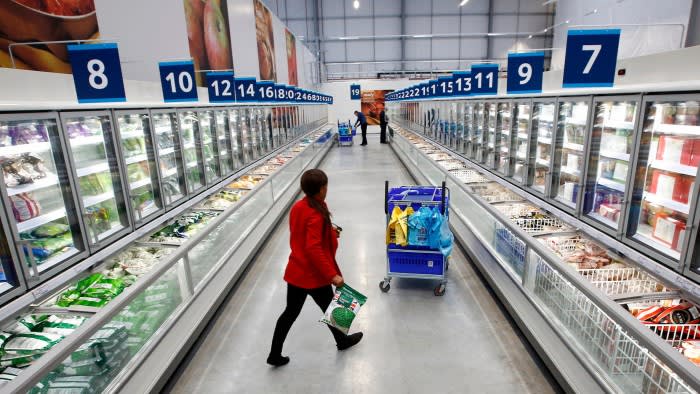 A Tesco employee in a red jacket carries a bag of frozen peas while walking through an aisle lined with large freezers. The aisle has numbered signs above the freezers, and a blue cart filled with plastic bags is visible in the foreground