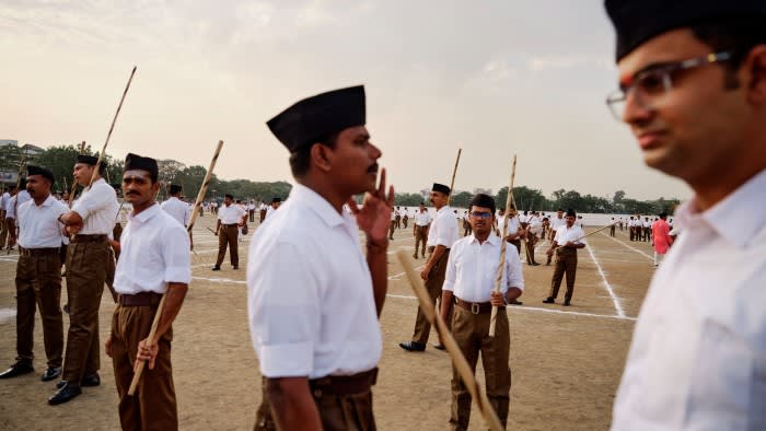 Men clad in brown trousers, crisp white shirts and black caps in an open field outdoors. They carry bamboo sticks