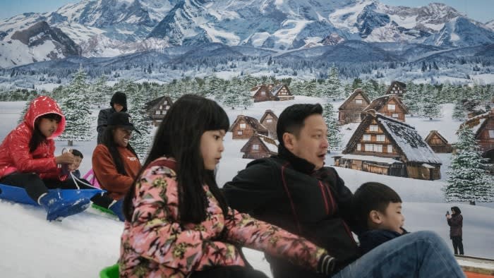 People ride sleighs on artificial snow at an indoor snow park with a mural of snowy mountains and traditional houses in the background