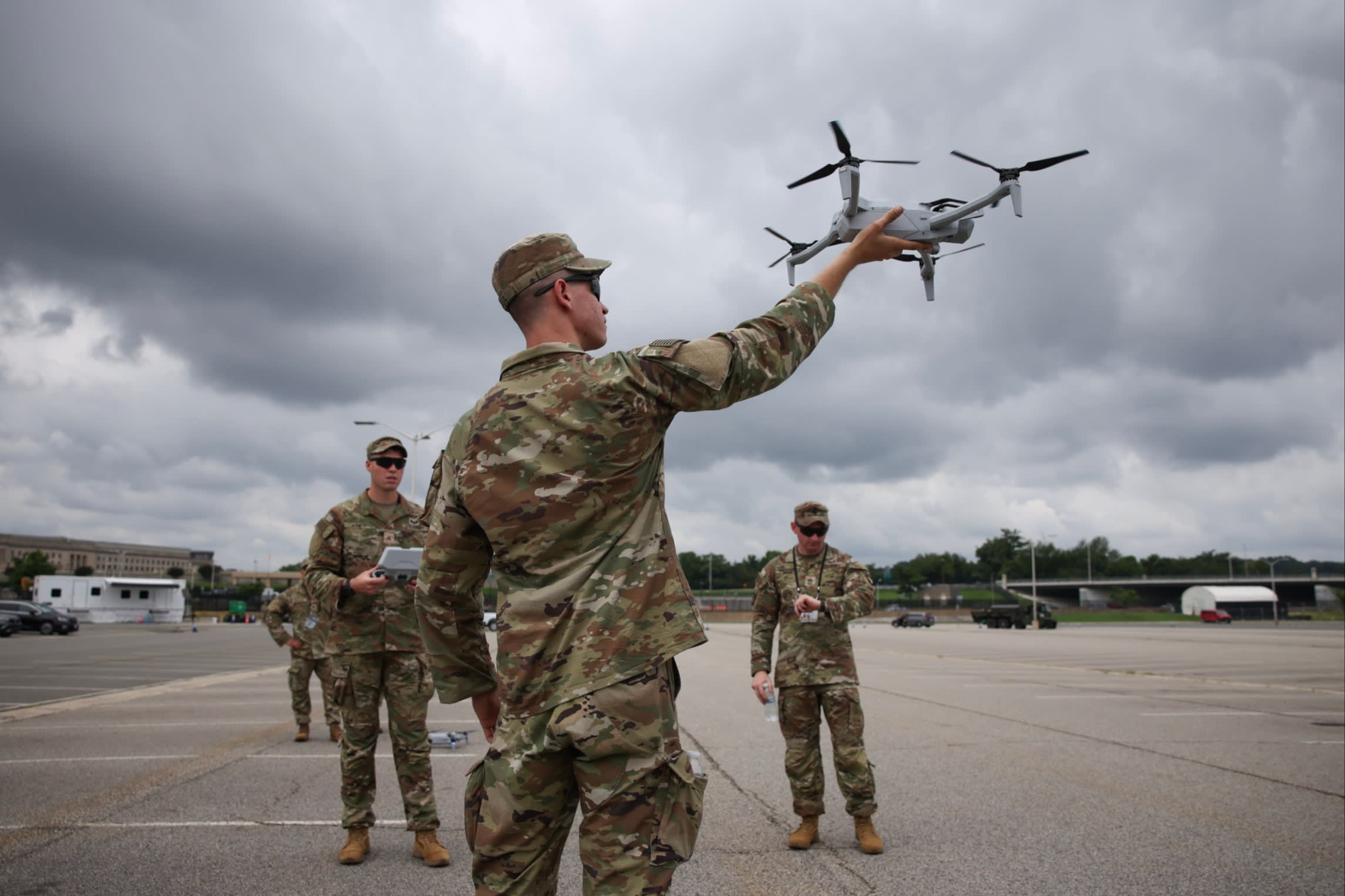 A US soldier holds a drone aloft in the Pentagon parking lot, with other soldiers standing nearby under a cloudy sky.