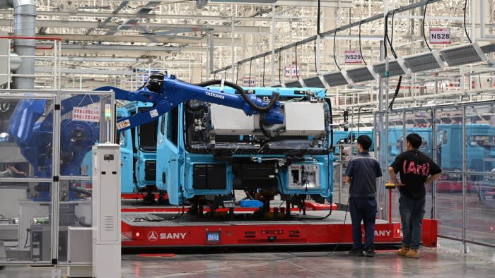 Blue electric truck cabs on an assembly line at the Sany factory, with two workers and a robotic arm operating nearby.