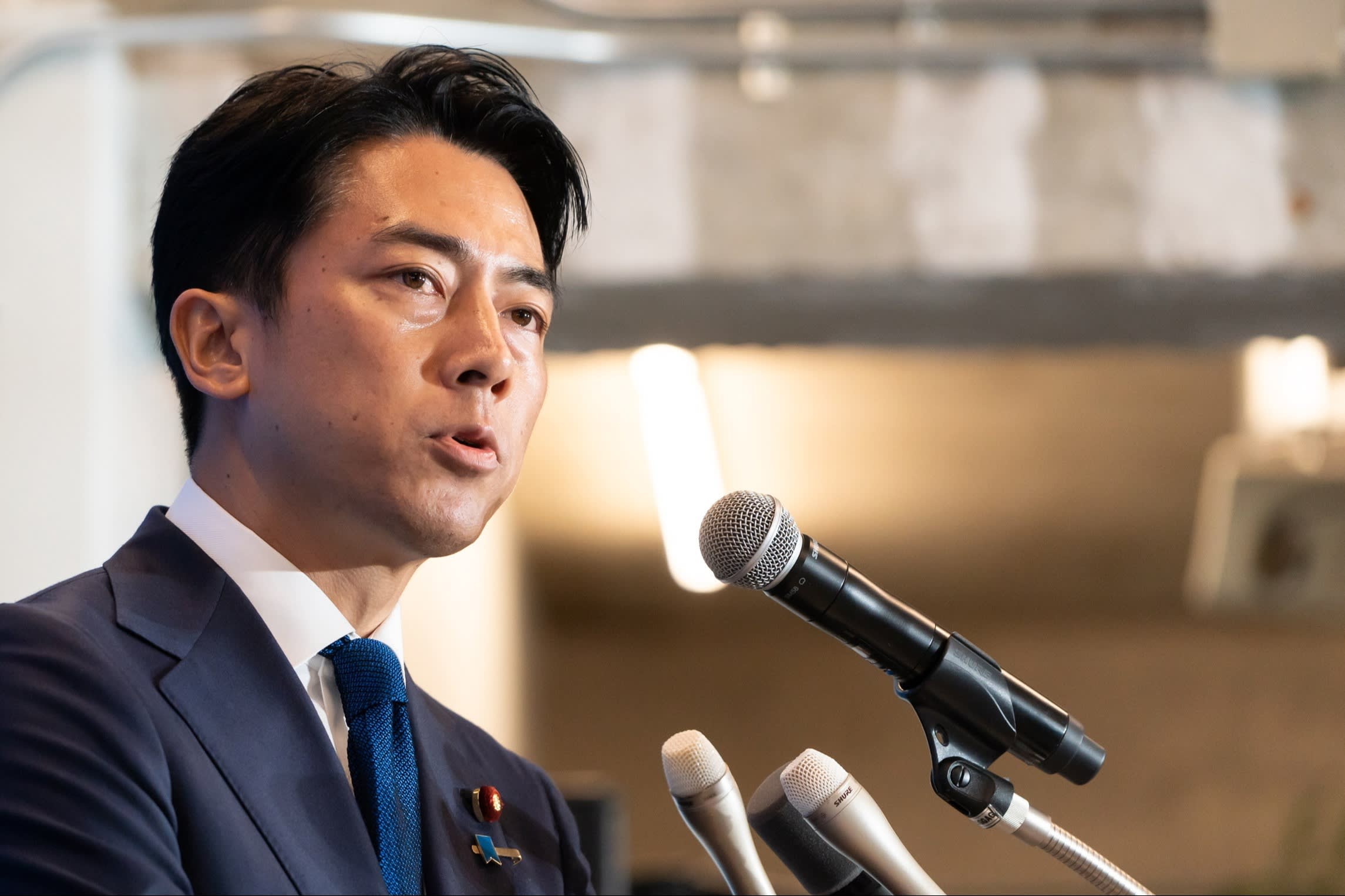 Shinjiro Koizumi speaks at a press conference, standing behind three microphones and wearing a suit and blue tie.
