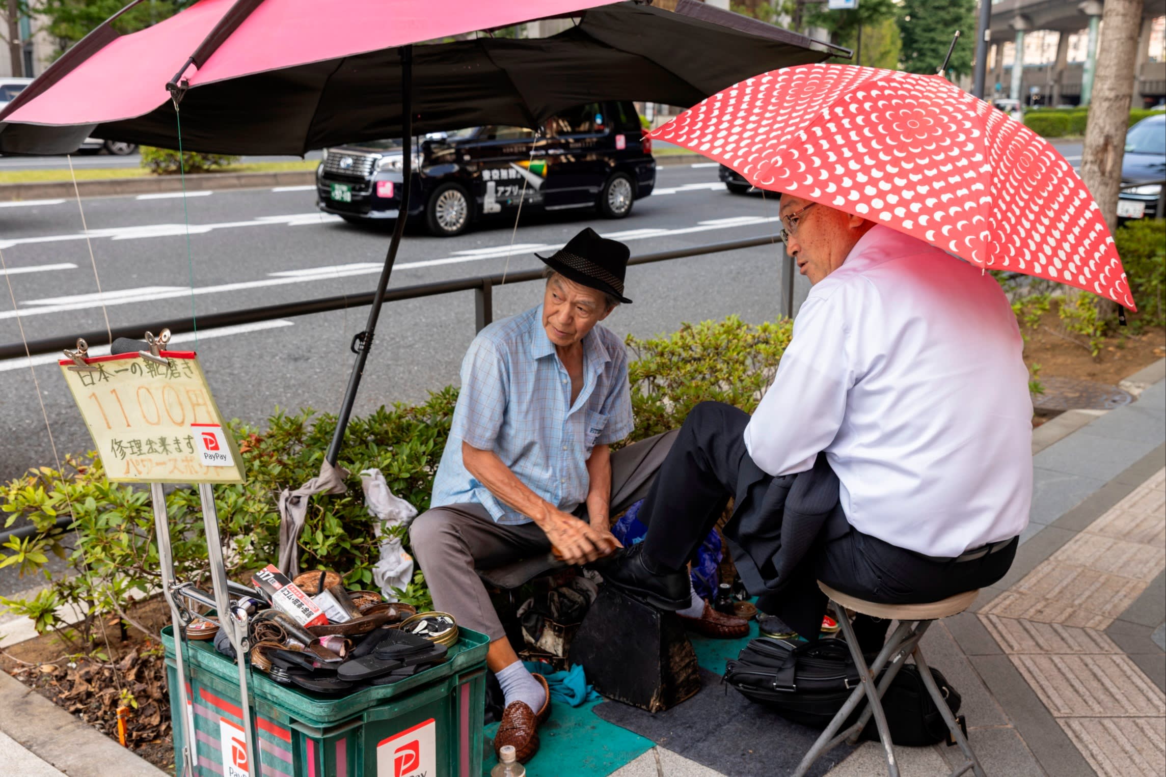 A shoeshiner polishes a customer’s shoe under umbrellas on a Tokyo sidewalk 