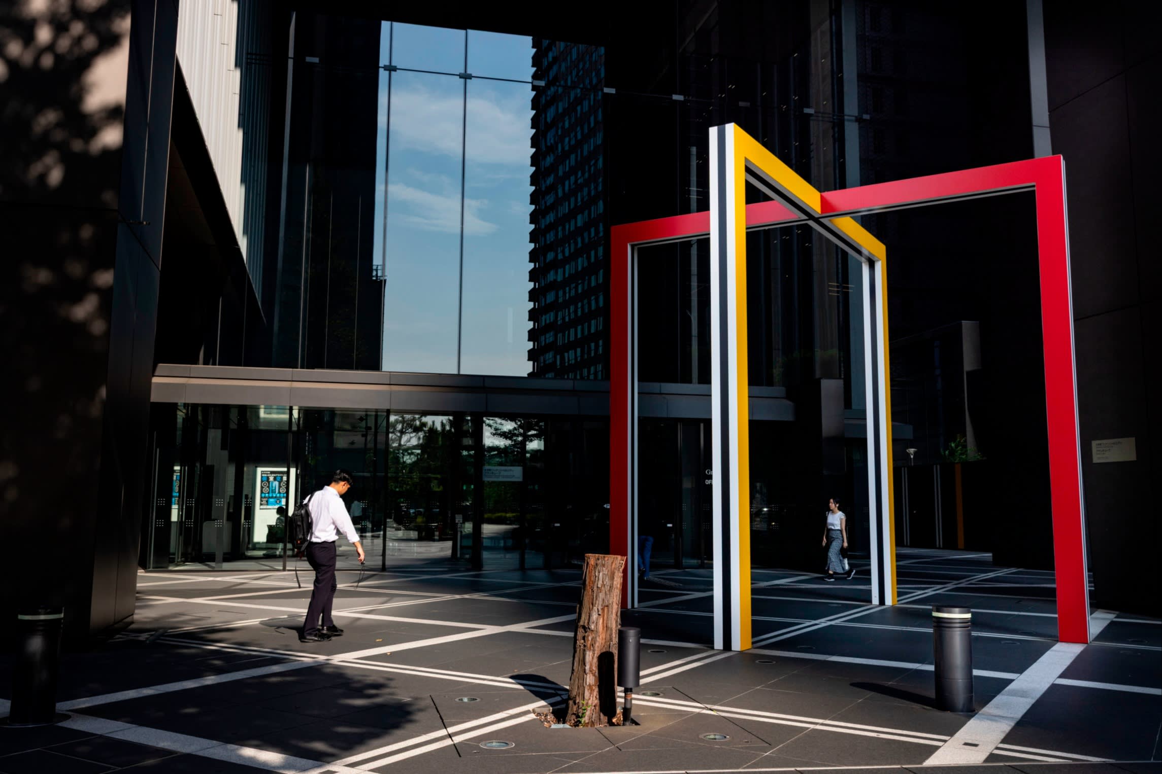 A man and a woman walk outside the Otemachi Financial City Grand Cube building, passing a large geometric sculpture with red and yellow frames.