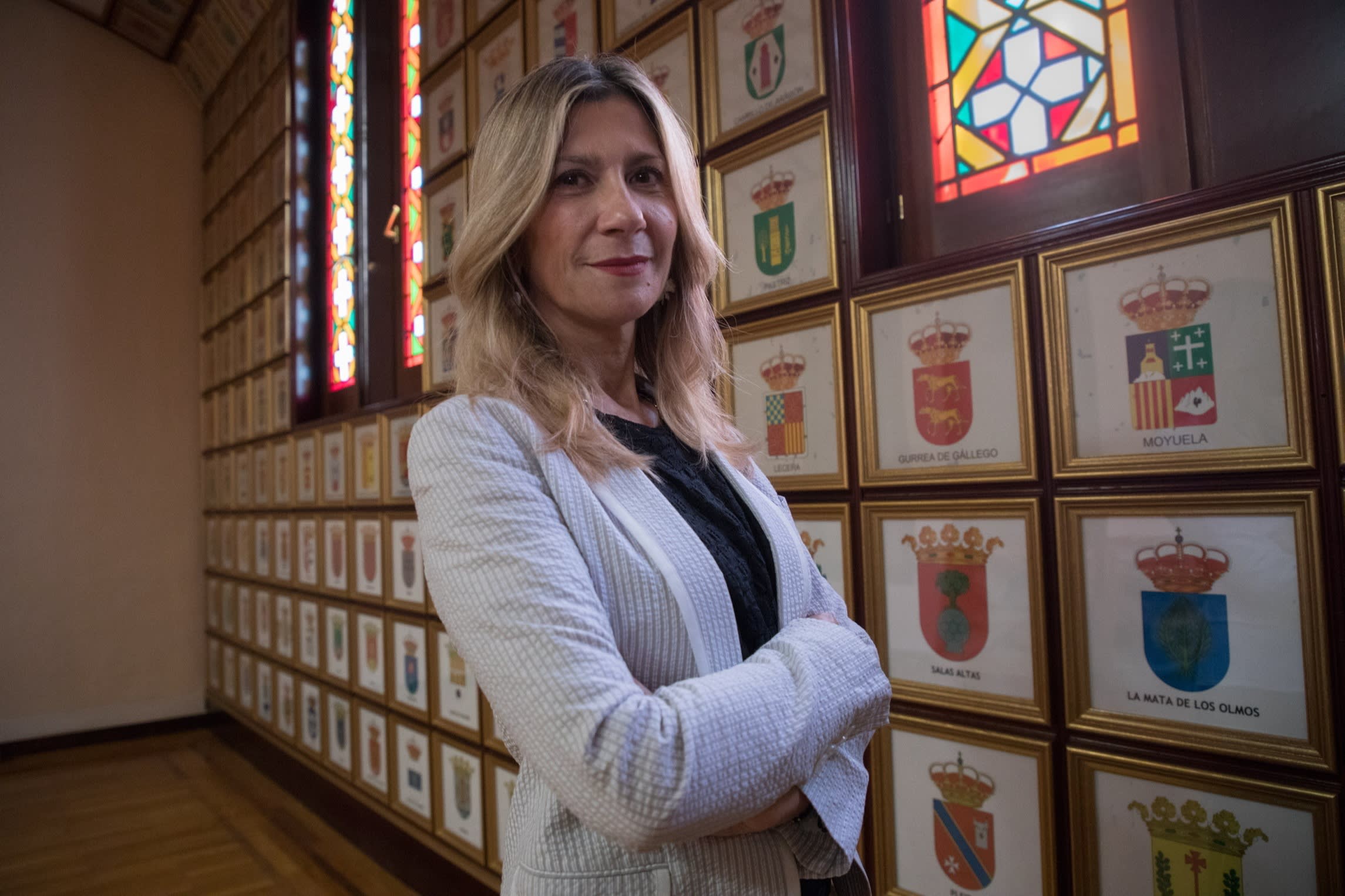 Mar Vaquero stands with arms crossed in the Coat of Arms Room, surrounded by framed municipal crests and stained glass windows.