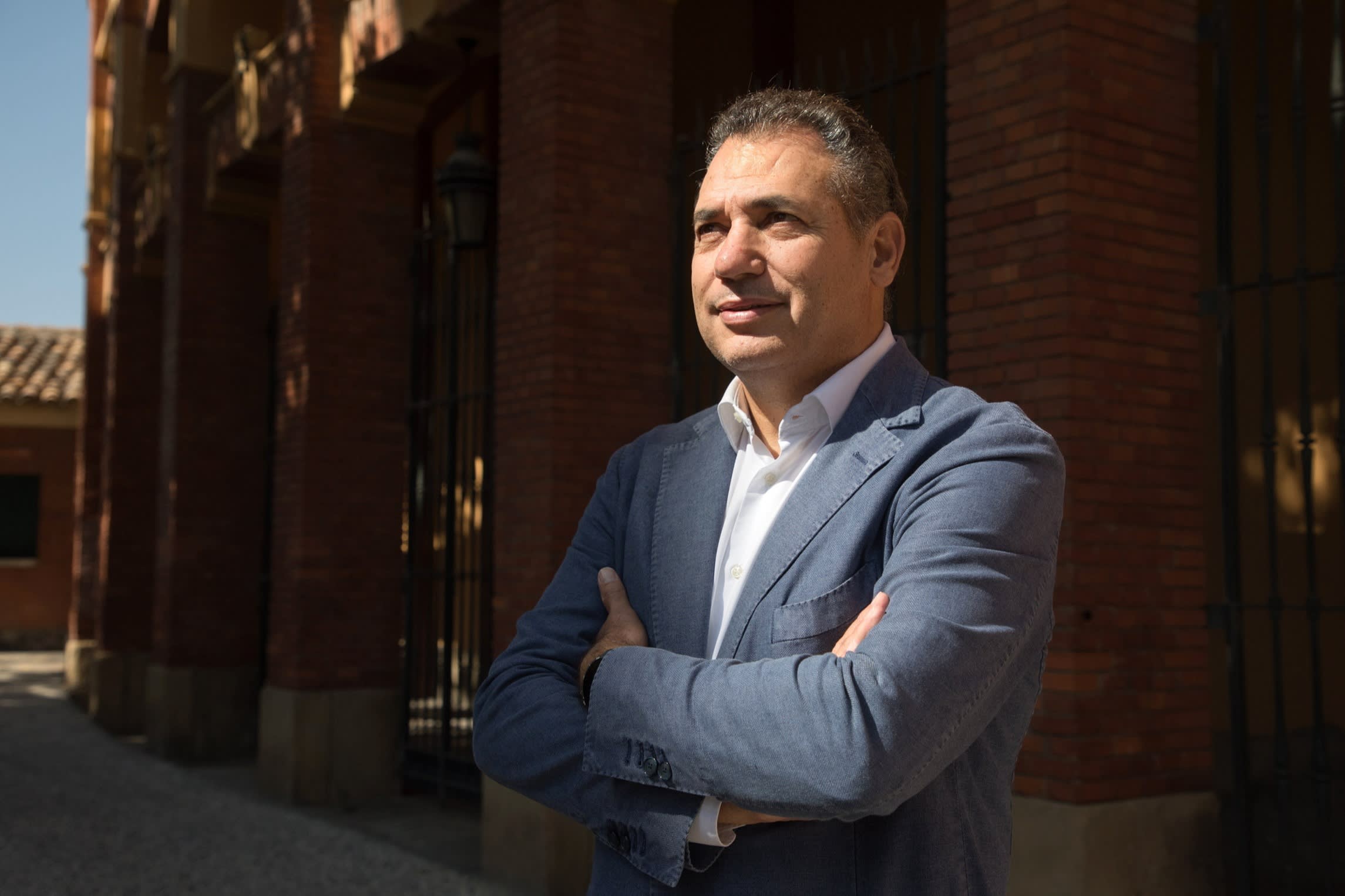 Benito Tesier Sierra standing outdoors with arms crossed, wearing a blue blazer and white shirt, in front of a brick building.