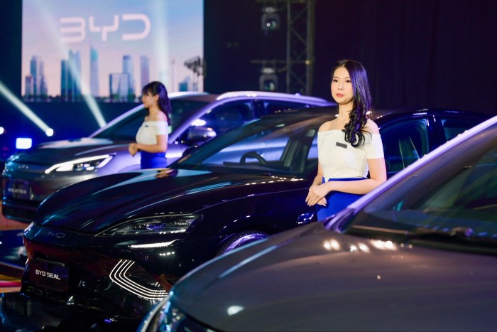 Models stand between three BYD electric vehicles at a launch event in Jakarta, with a BYD logo displayed on a screen in the background