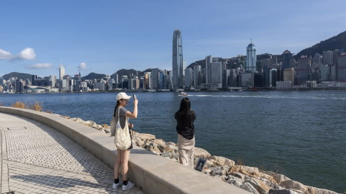 Two people take photos of the Hong Kong skyline from the West Kowloon waterfront on a sunny day