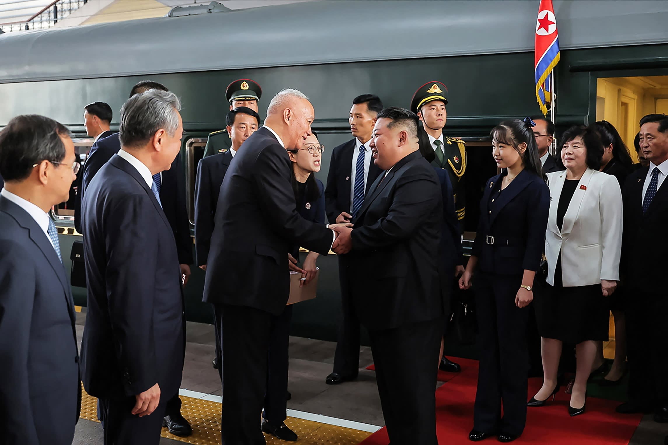 Kim Jong Un is greeted by Cai Qi at Beijing railway station, with Kim Ju Ae and Wang Yi nearby. Officials and guards stand in the background