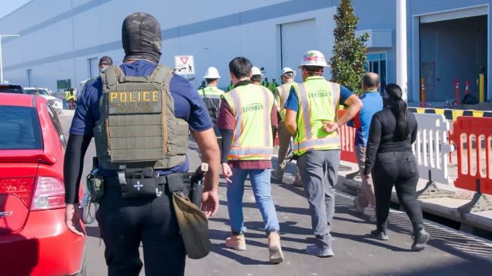 A Homeland Security Investigations police officer escorts a group of construction workers wearing safety vests and hard hats outside an industrial building.