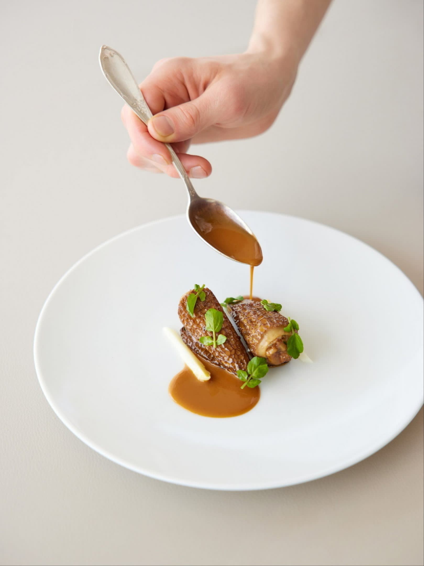 A hand pours sauce from a spoon onto an artfully plated dish with morel mushrooms stuffed with Botan shrimp and garnished with herbs at Sézanne.