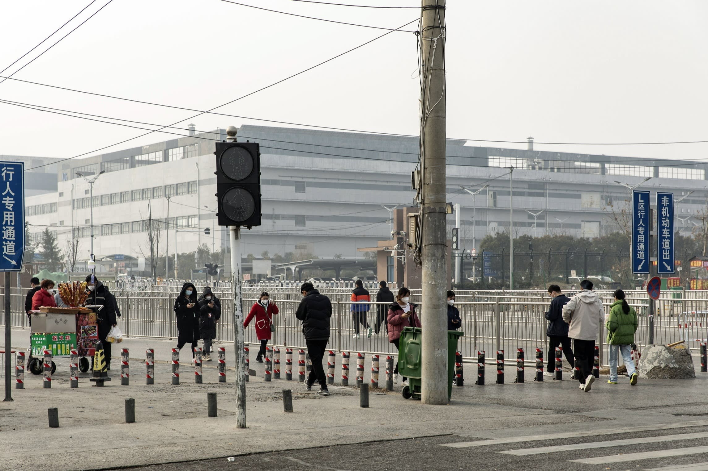 People walk near a large factory building, some wearing masks, with a street vendor cart and traffic signs visible in the foreground.