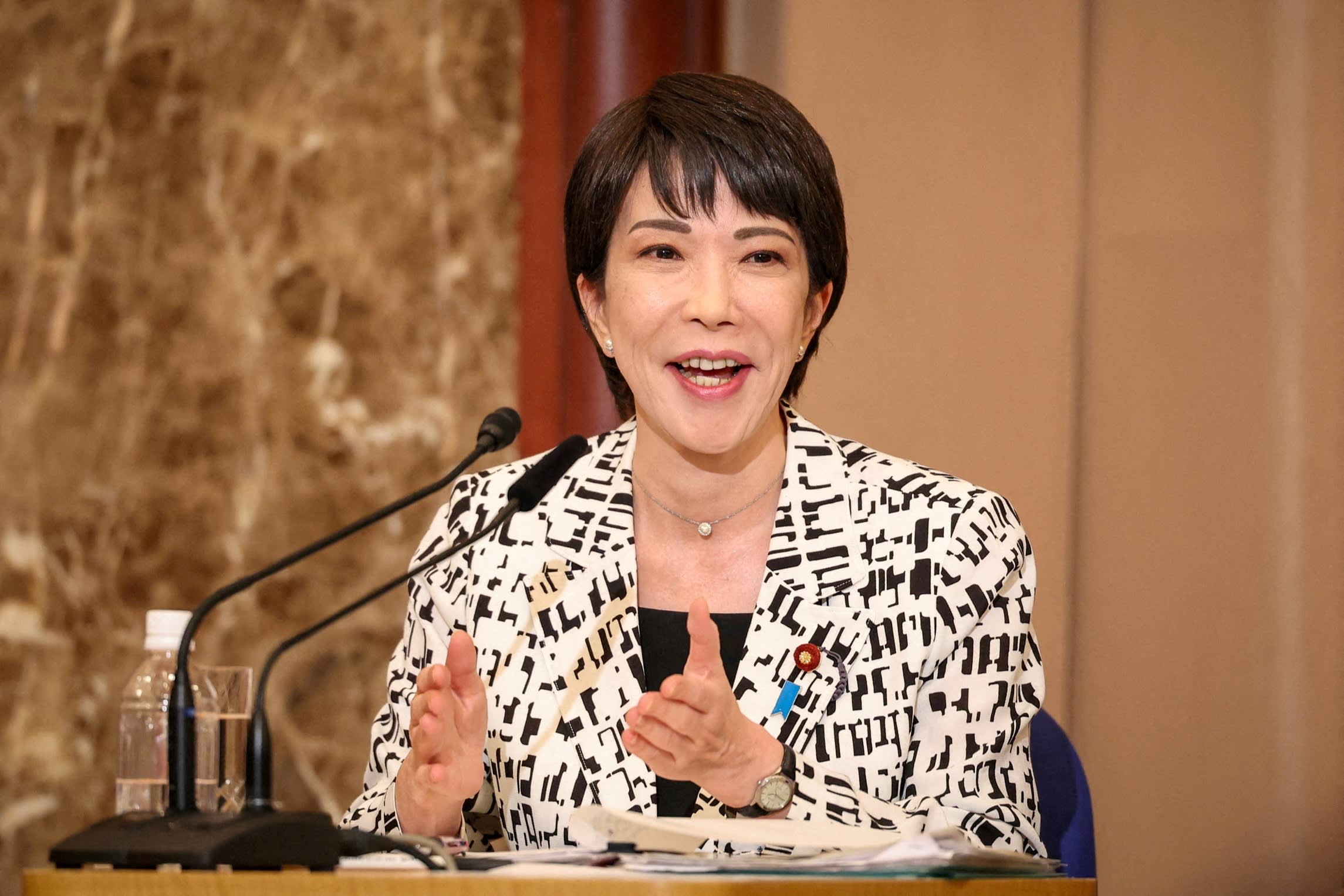 Sanae Takaichi speaks into a microphone during a debate, gesturing with her hands and smiling.