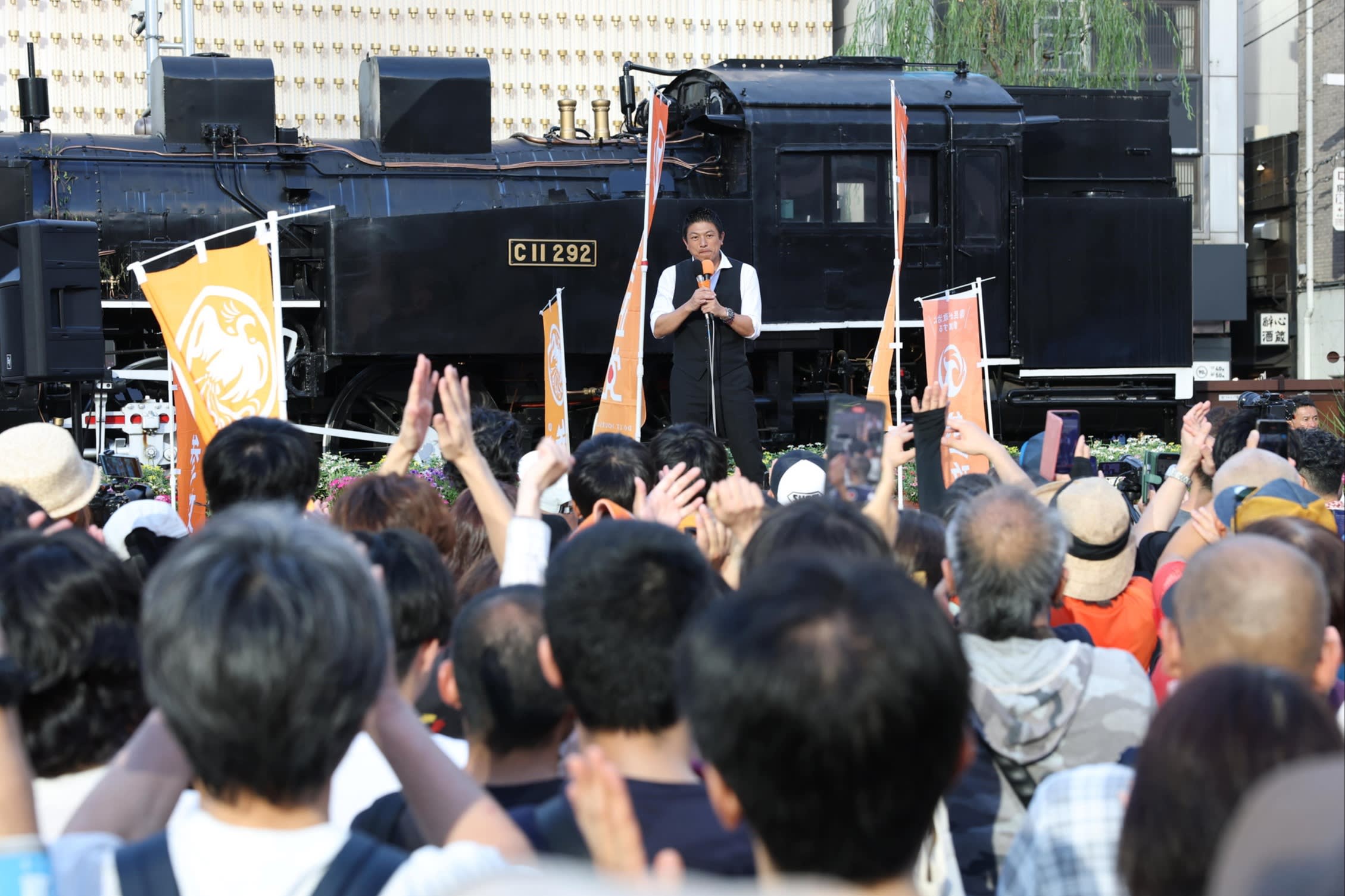 Sohei Kamiya speaks to a crowd during a street speech in front of a steam locomotive, with supporters raising their hands.