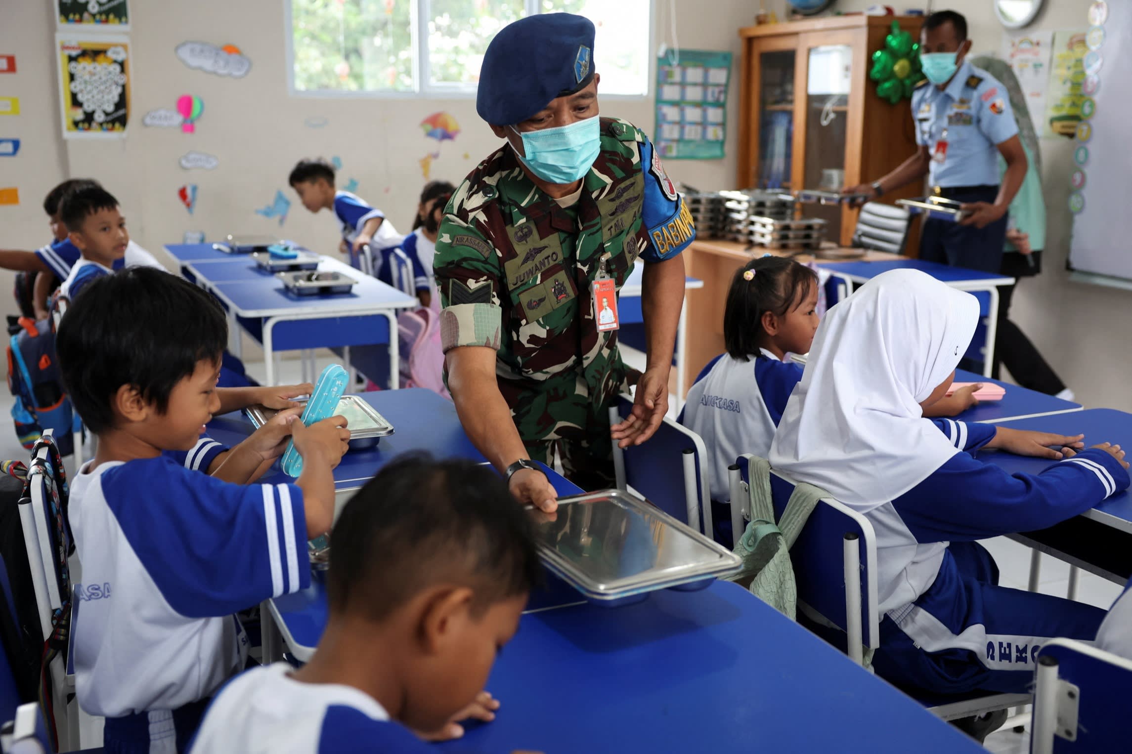 Juwanto, an Indonesian Air Force personnel, hands a meal box to a student seated in a classroom with other children at Angkasa 5 elementary school.