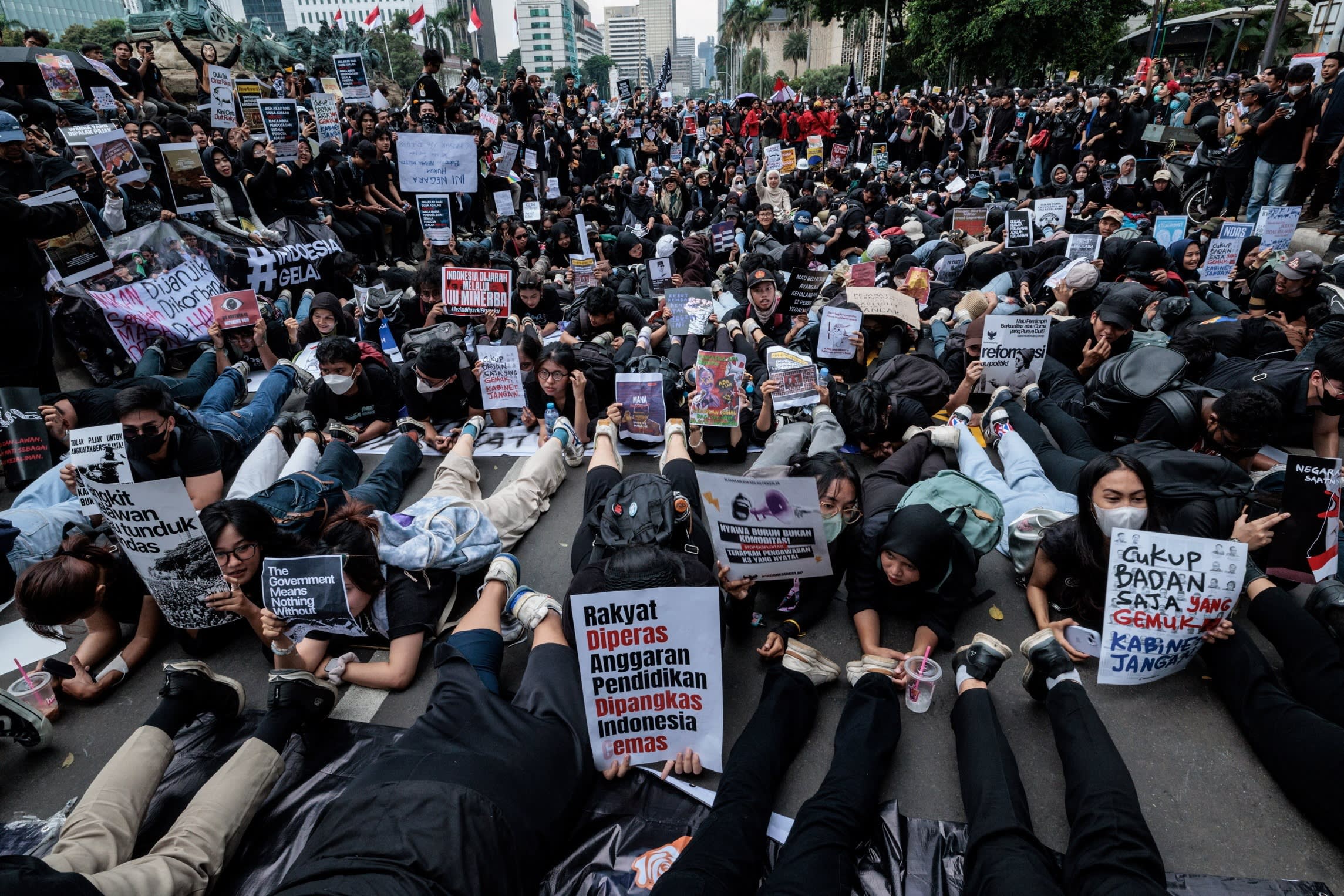 Hundreds of protesters sit on a city street, holding signs with demands and slogans critical of government policies.