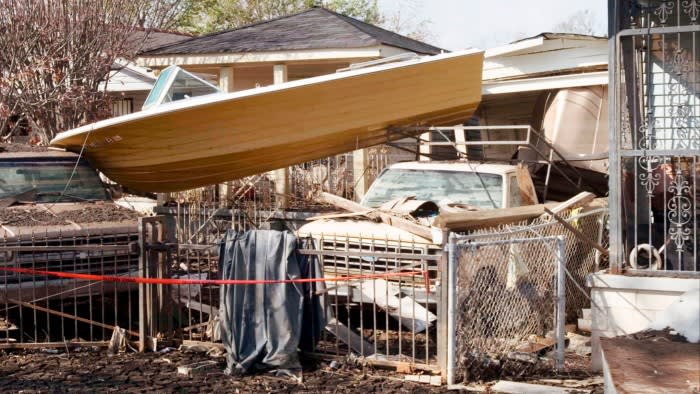 A speedboat perched on top of two pick-up trucks in front of a hurricane-wrecked house