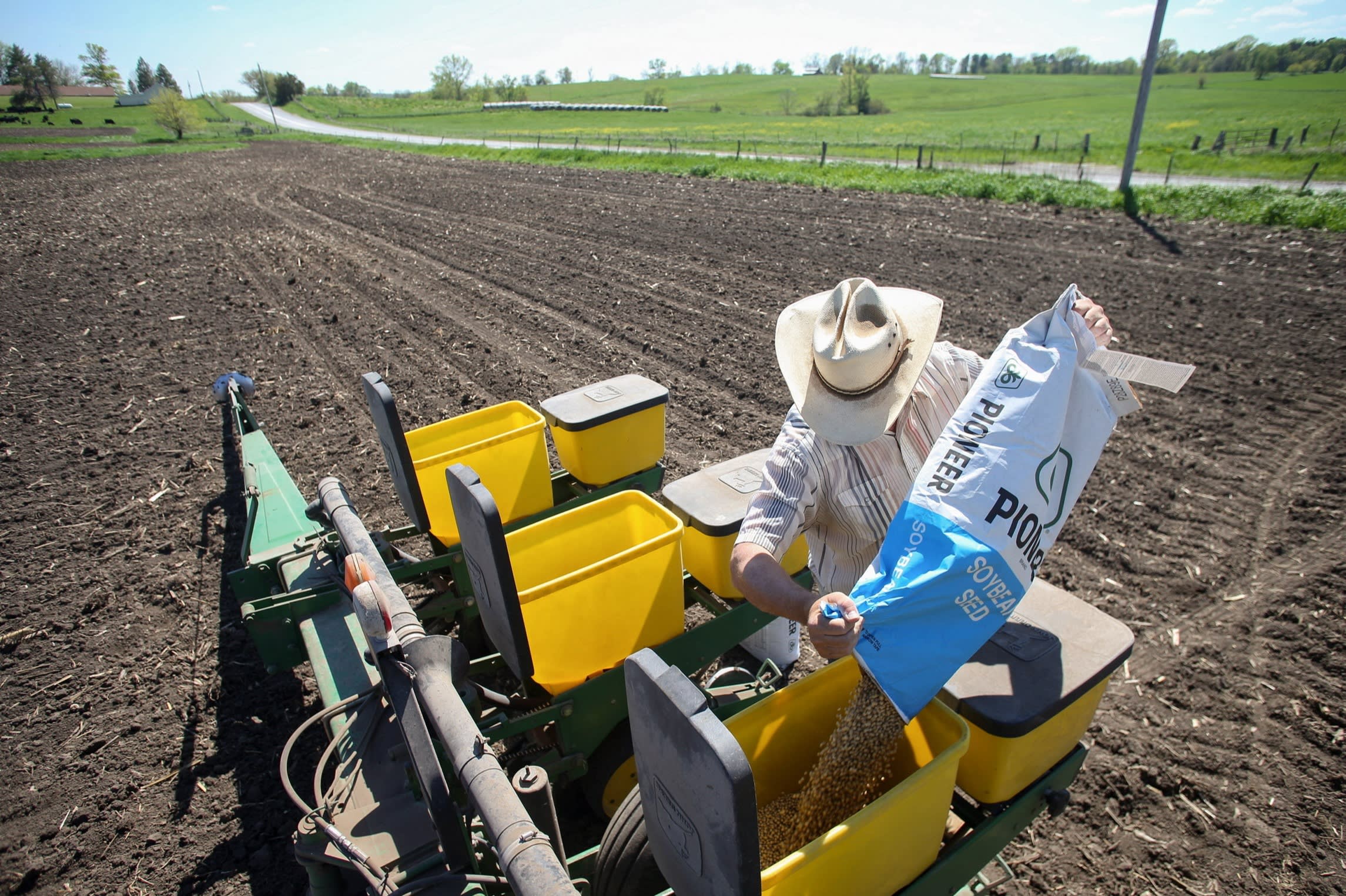 A worker in a cowboy hat pours soyabean seeds from a large Pioneer bag into a planter on a freshly tilled field.