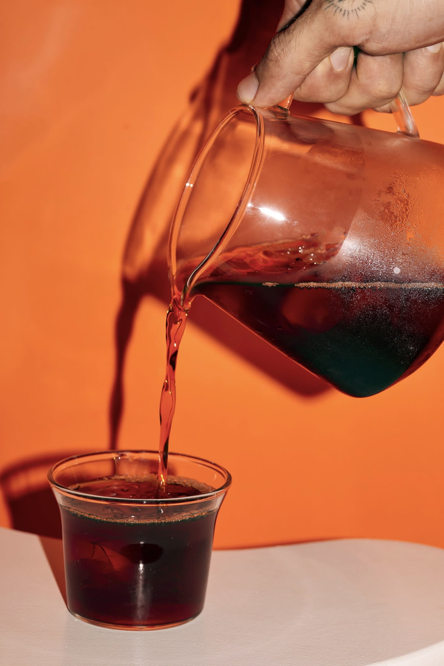 A hand pours black coffee from a glass pitcher into a glass cup against an orange background at Café Nuances.