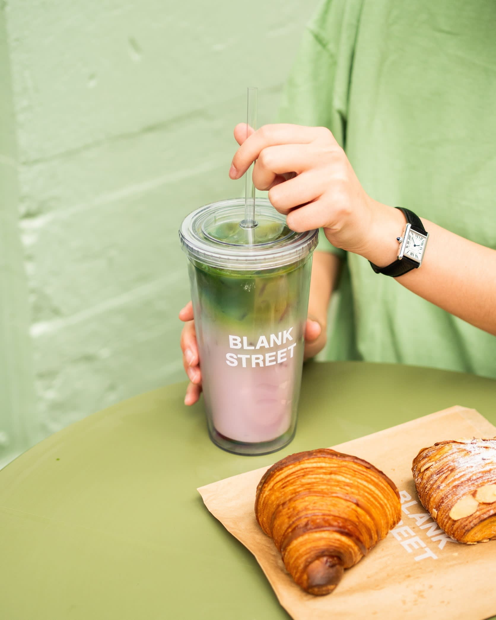 A person in a green shirt holds a cup labeled &quot;Blank Street&quot; with a layered blueberry matcha drink. Two pastries are on a paper bag on the table.