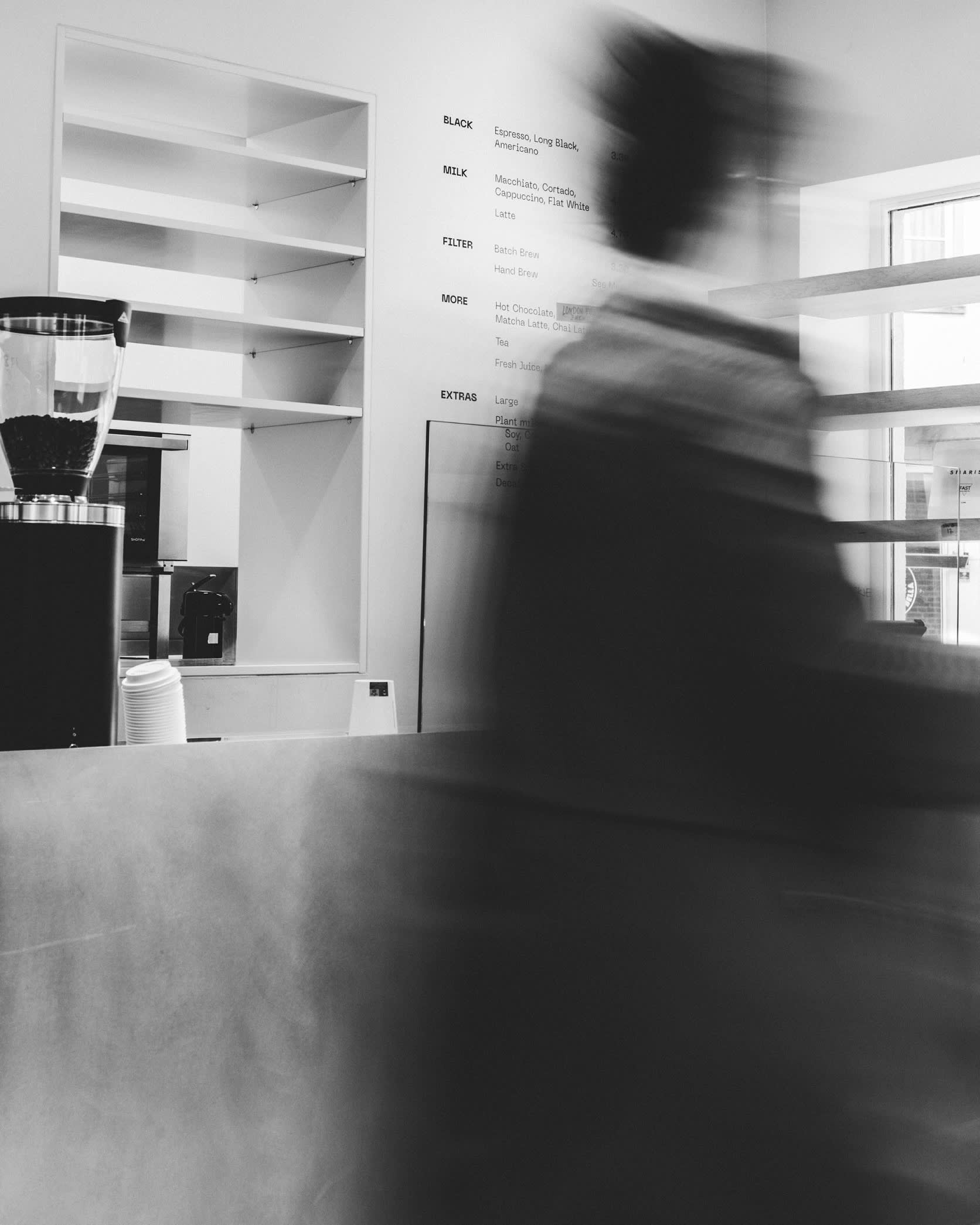 A blurred figure moves past a minimalist coffee shop counter with empty shelves and a visible menu on the wall at the outpost of Bouche in Brussels that is near the EU institutions.
