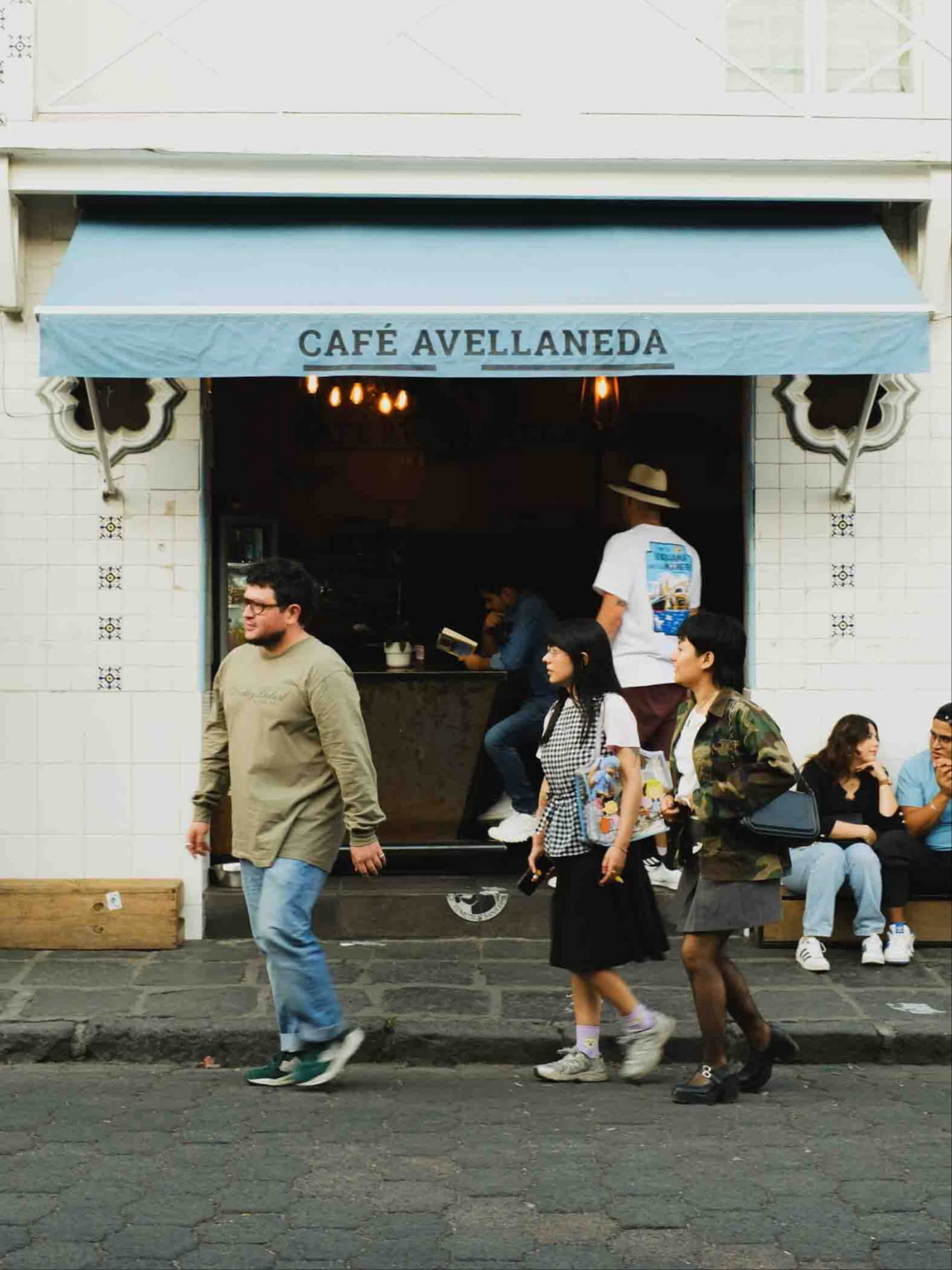 People walking by the white façade of Café Avellaneda in Mexico City. Above the door is a pale-blue canopy on which Café Avellaneda is printed in black lettering 