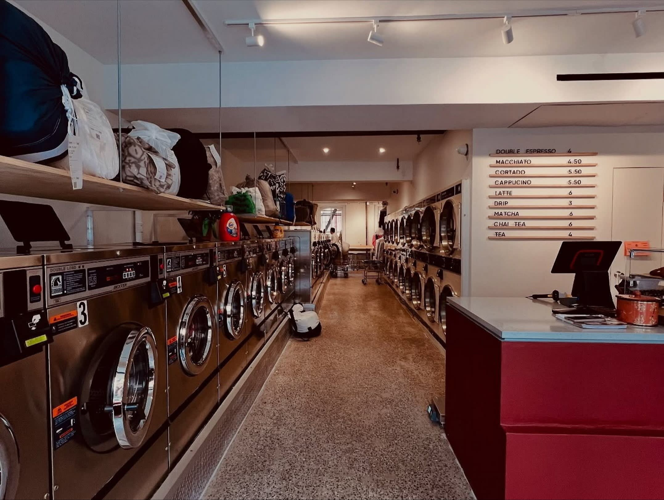 A row of industrial washing machines lines one wall of Laundry & Latte in Brooklyn, with shelves above holding bags of laundry. On the opposite wall is a menu board listing coffee drinks and prices, and a counter with a register is in the foreground.