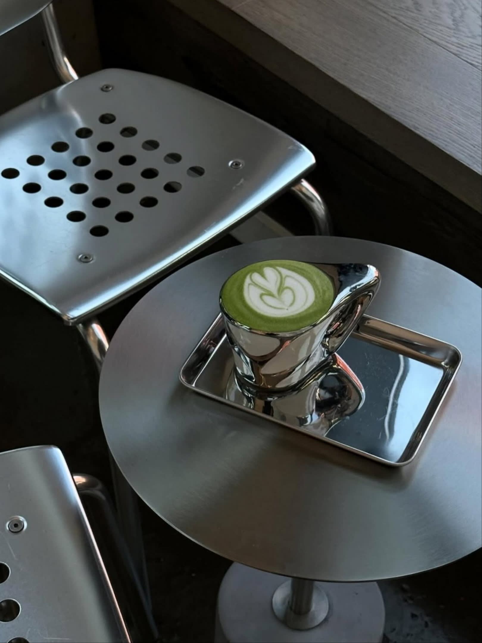 A shiny metallic cup with matcha latte art sits on a metal tray atop a round metal table, next to perforated metal chairs, at Café MSMN in Greenpoint.