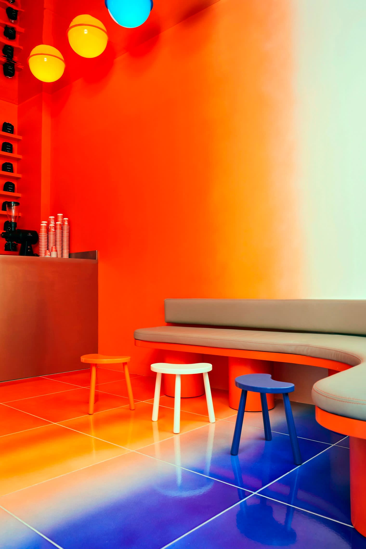 The colourful interior of Café Nuances in Paris, featuring orange walls, a gradient orange-to-blue tiled floor, small colourful stools and a curved bench.