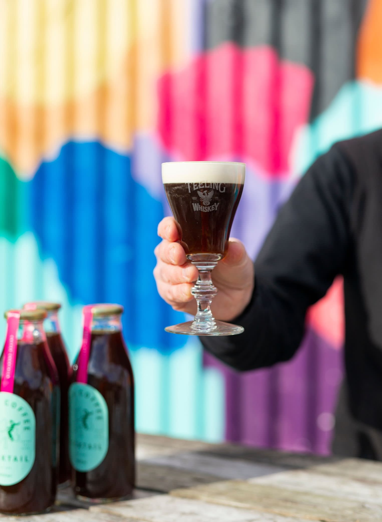 A person holds a glass of Irish coffee made with poitín with a creamy top layer at Vice Coffee Inc in Dublin, with bottles of coffee cocktail on a table and a colourful mural in the background.