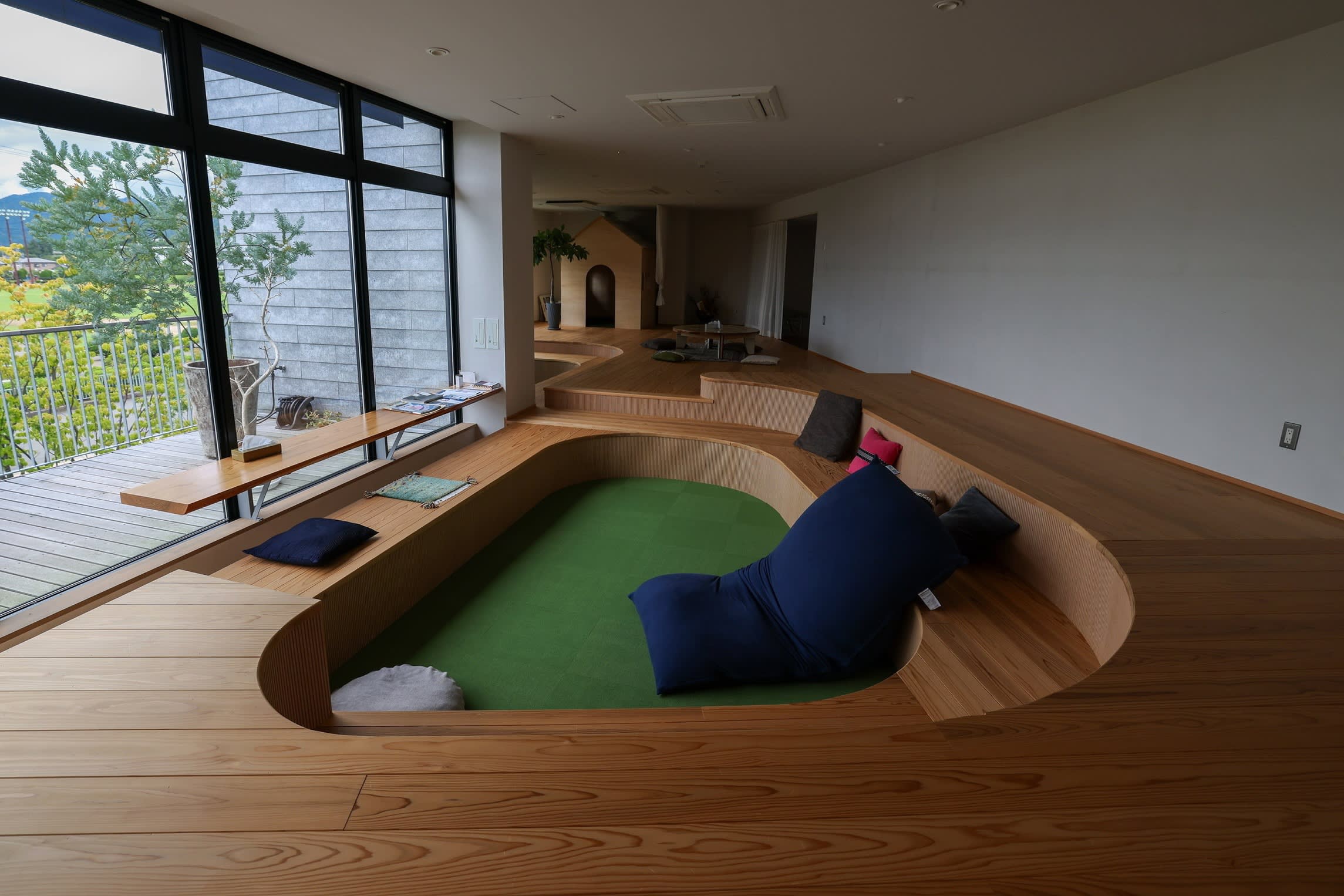 A lounge with sunken seating, colorful cushions, and large windows overlooking an outdoor view at Matsukawa Rapyarn.